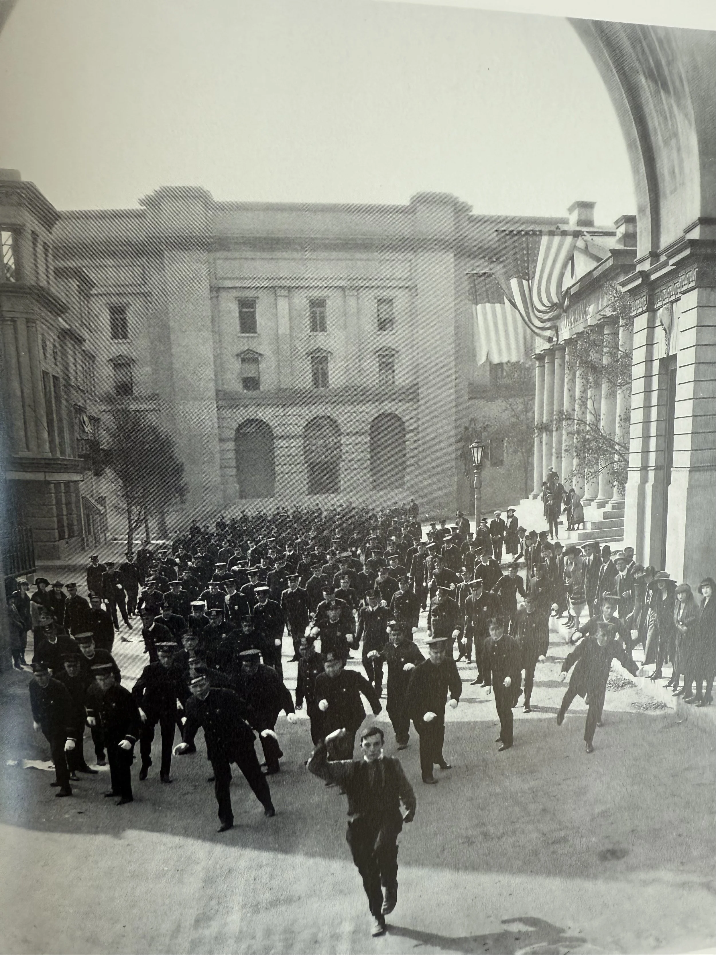Historical black and white photograph showing a large crowd of people walking outside a government building with tall columns, some carrying umbrellas, and American flags hanging from the facade.