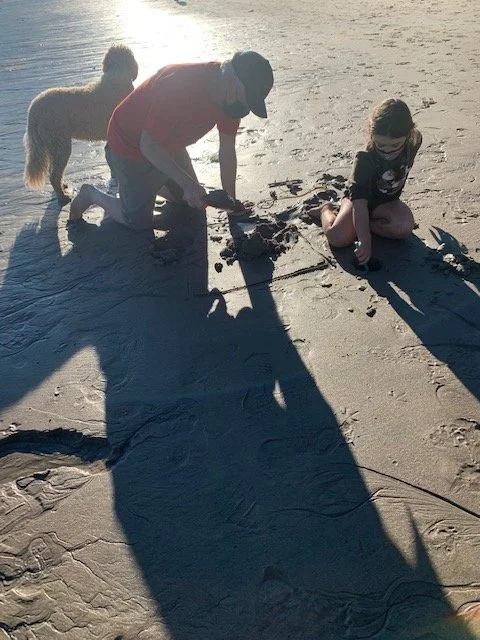A man, a girl, and a dog at the beach. The man and girl are drawing in the sand, with the man kneeling and the girl sitting. The dog is standing nearby. The setting sun casts long shadows on the sand.