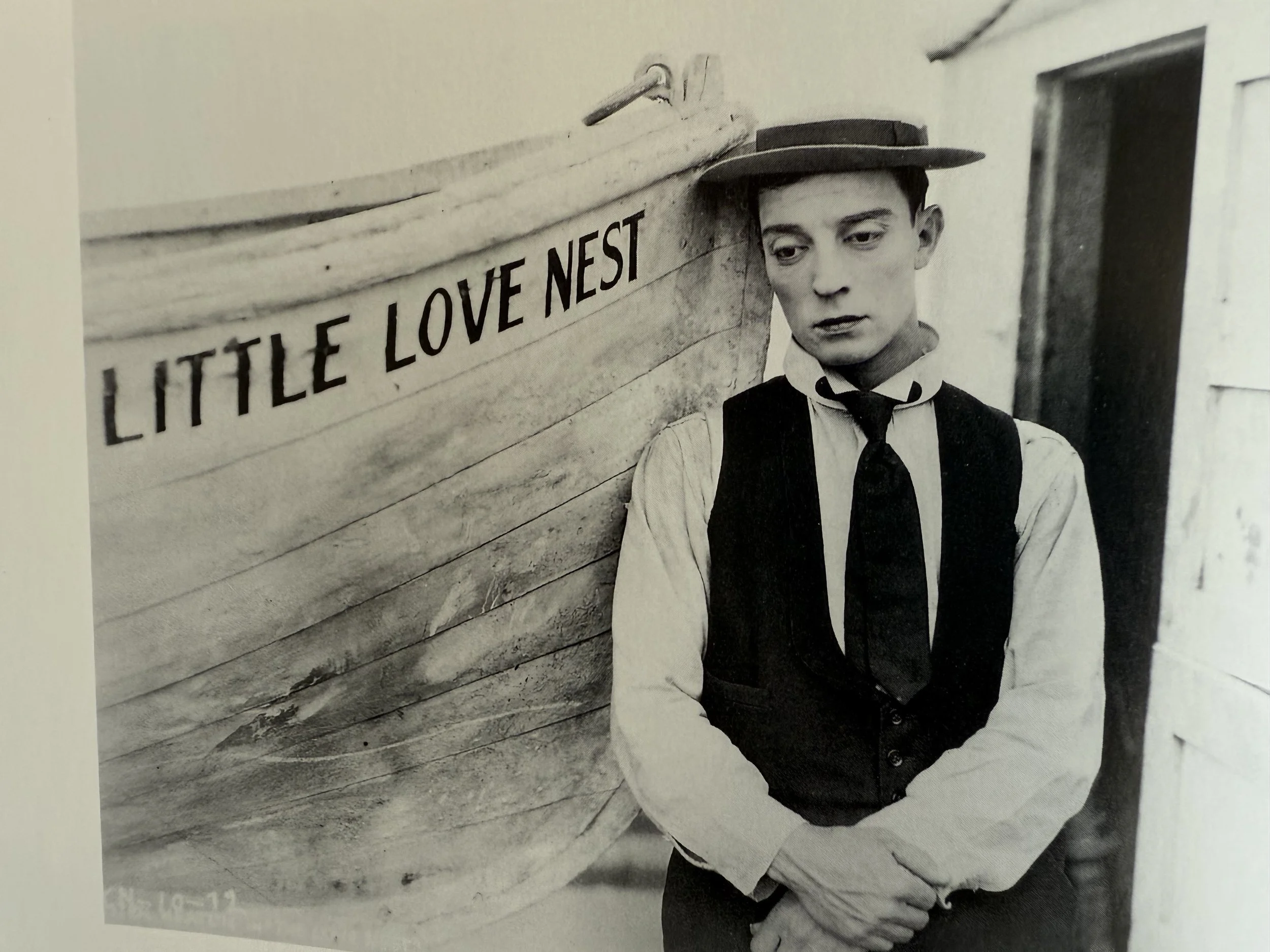 A black-and-white photo of a young man in vintage clothing, leaning against a wall with a sign that reads 'LITTLE LOVE NEST'. He wears a hat and a vest, with a serious expression.