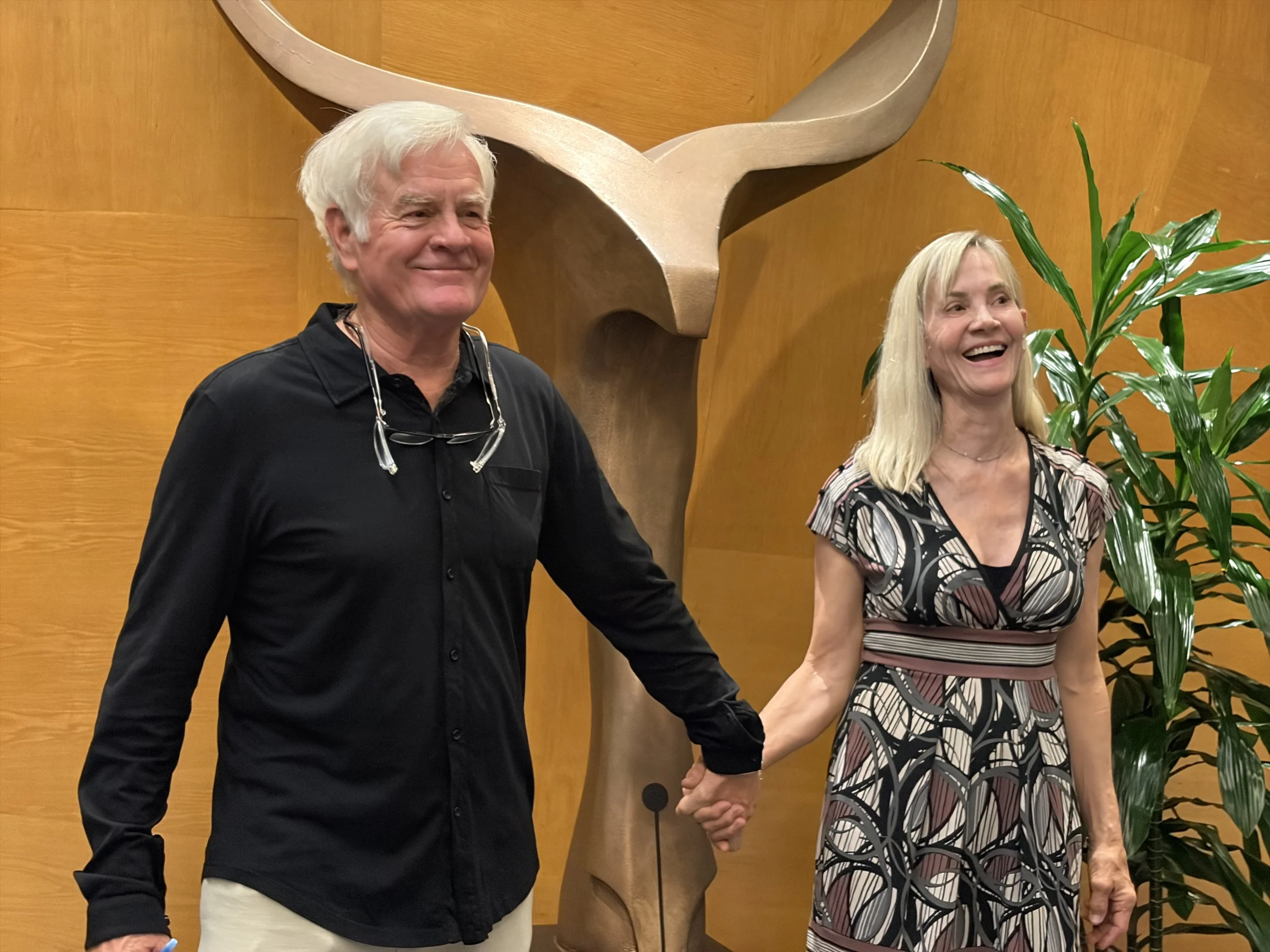 A man and woman holding hands and smiling in front of a wooden wall with a logo or sculpture, a large green plant on the right.