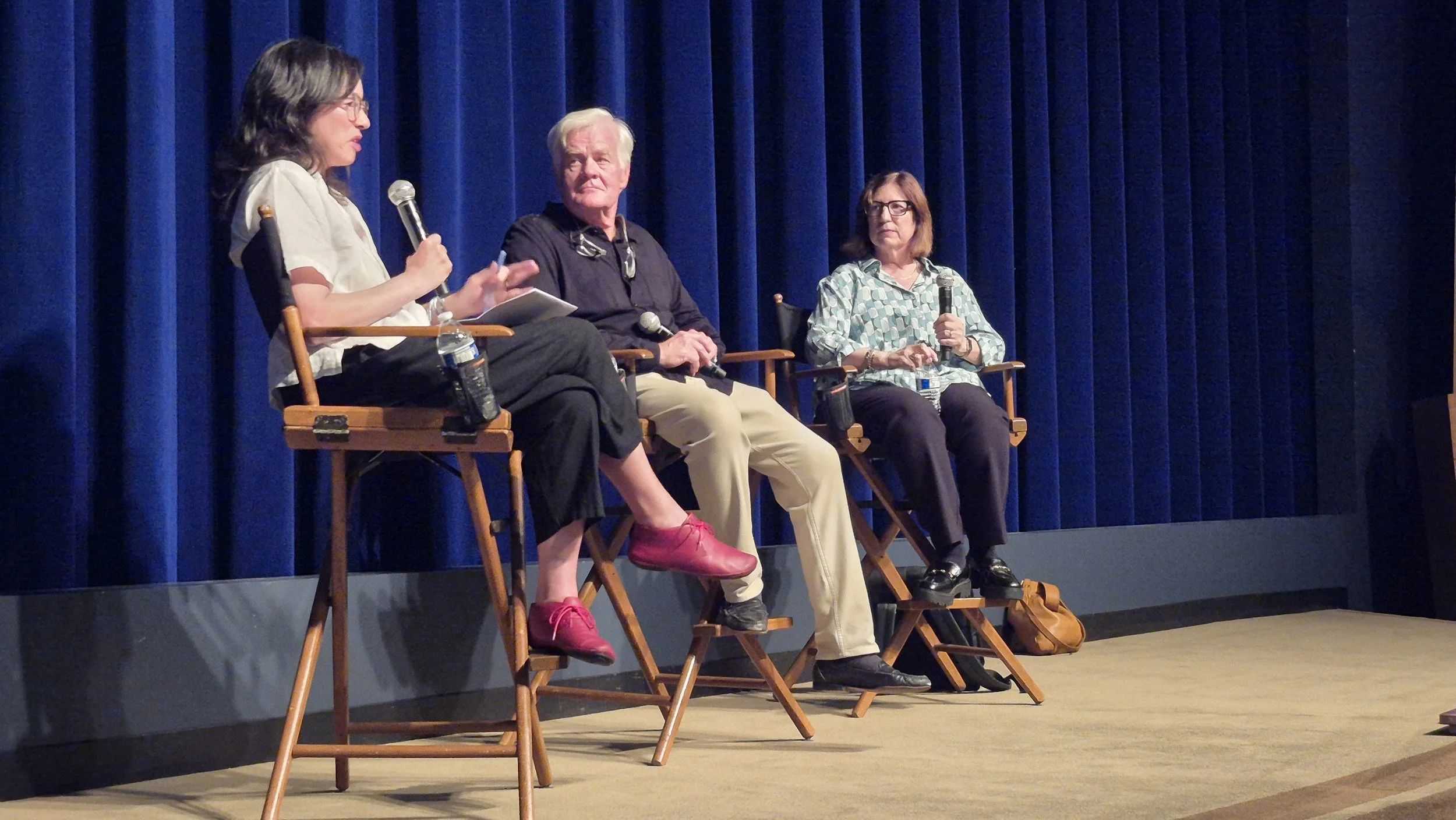 Four people sitting on chairs on stage with blue curtains in the background, participating in a panel discussion. The woman on the far left is speaking into a microphone, the man next to her is looking at her, and the other woman is holding a microph