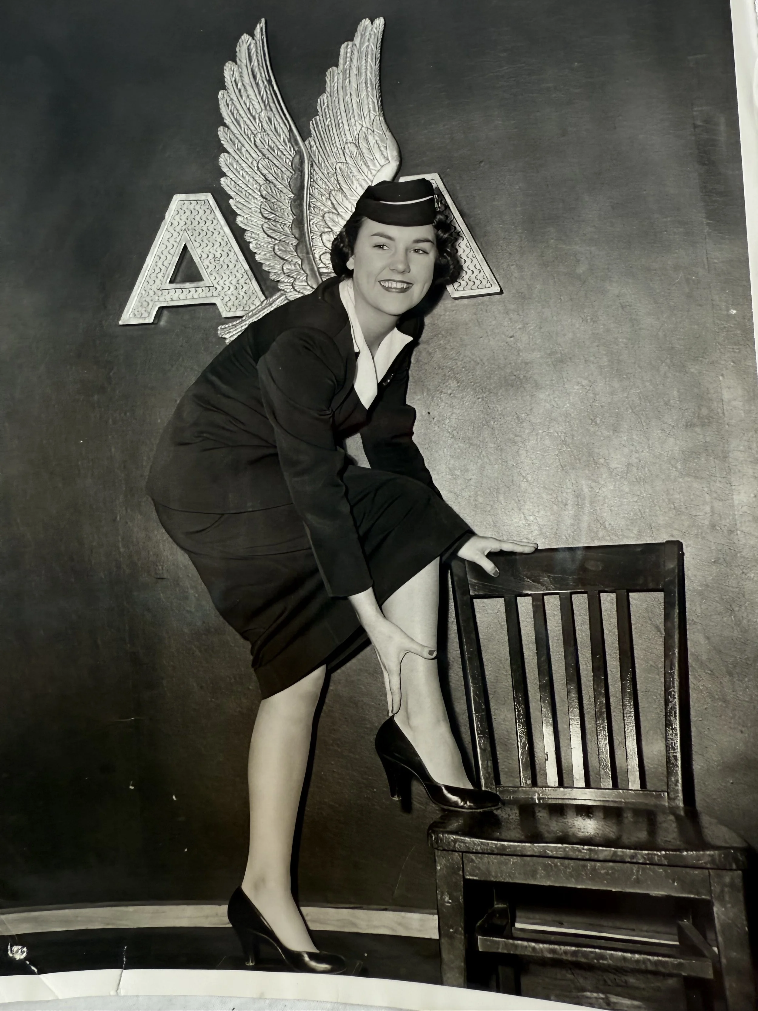 Black and white image of a woman in a nurse uniform with vintage style, wearing high heels, sitting on a wooden chair, with large decorative wings, and a letter 'A' in the background.