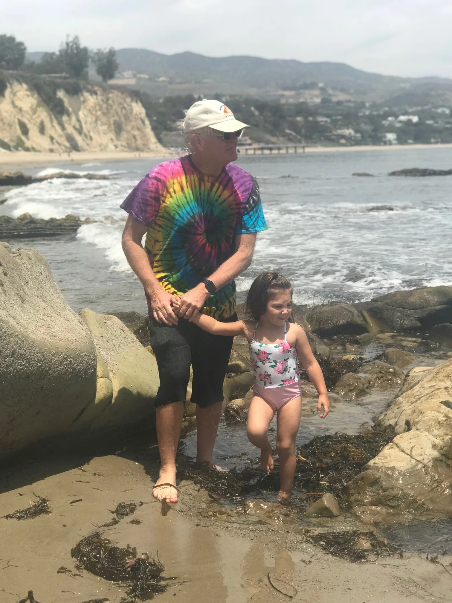 An elderly man wearing a colorful tie-dye shirt, cap, and sunglasses holds hands with a young girl wearing a floral swimsuit as they stand in shallow ocean water near rocks on a beach, with cliffs and houses in the background on a cloudy day.