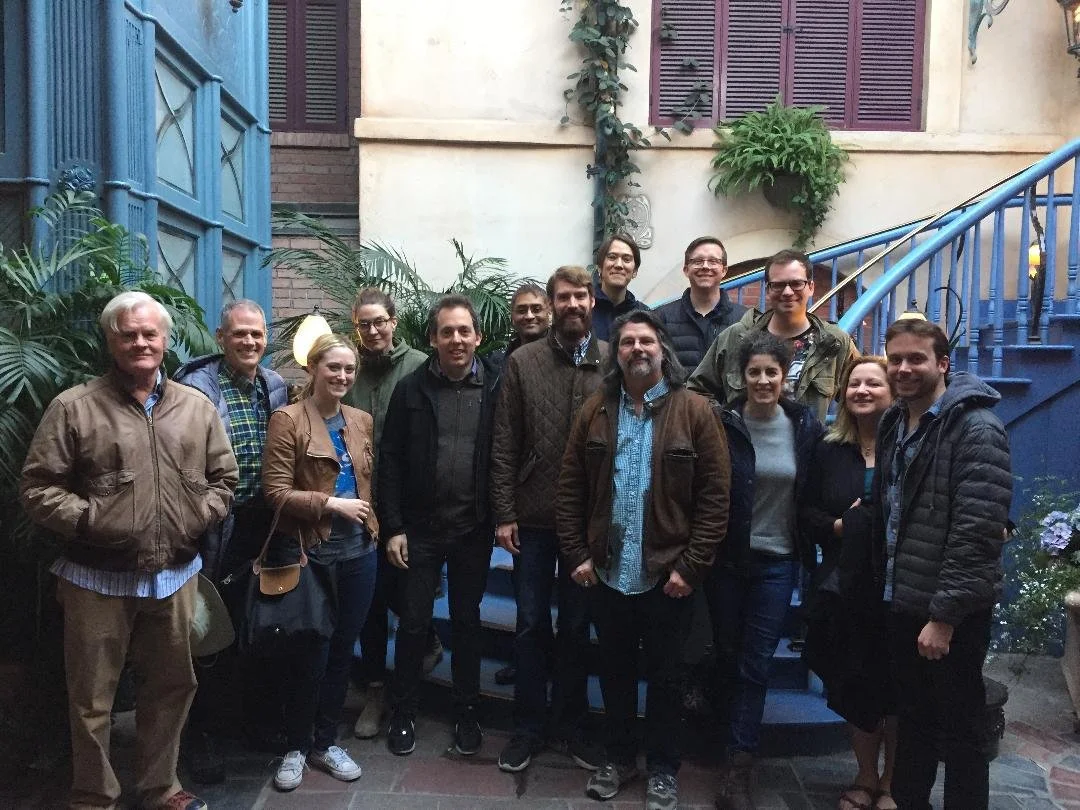 Group of fifteen people standing in a semi-circle indoors, with a blue staircase and tropical plants in the background, smiling for the photo.