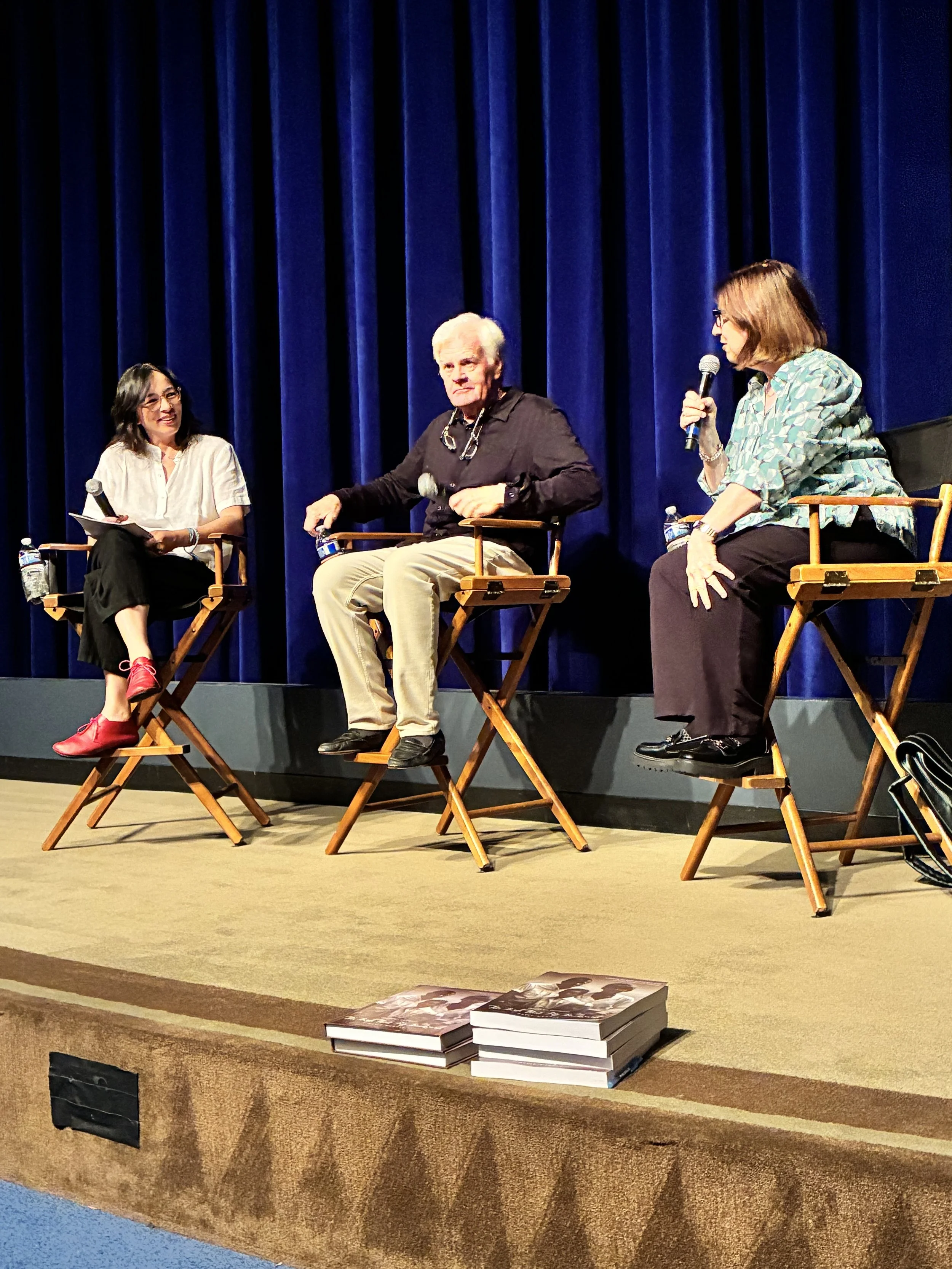 Three women engaged in a panel discussion on stage with blue curtains in the background. Two women are seated on wooden chairs, one holding a microphone, and a third woman stands on the left holding papers. There are books on the stage floor in front