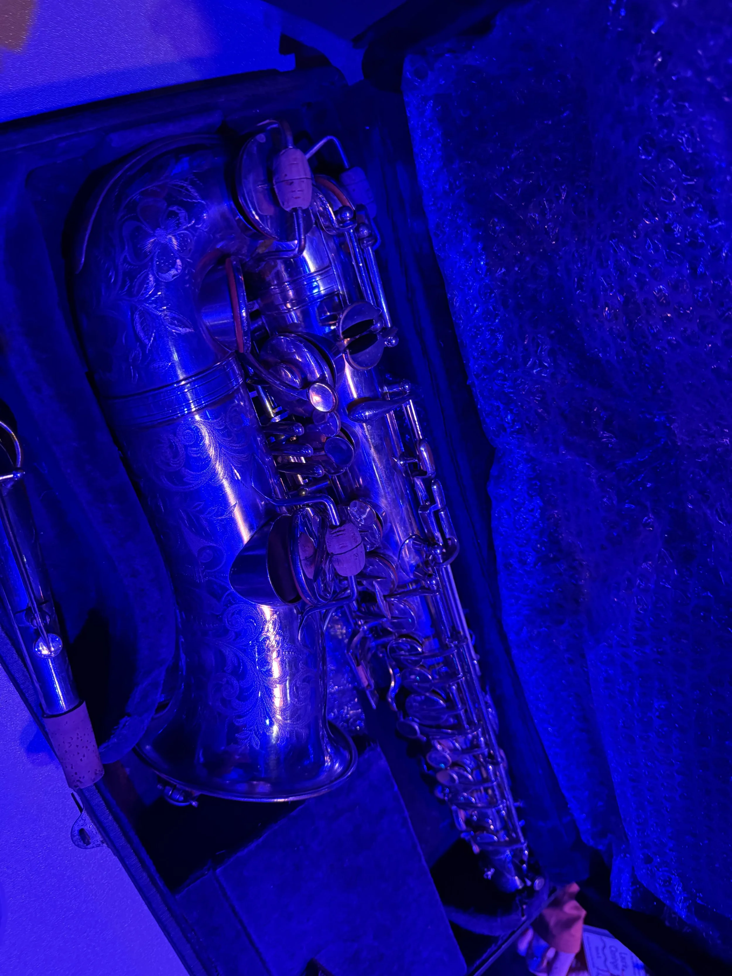 Close-up view of a silver saxophone with engraved floral patterns, illuminated by blue light, resting on a surface next to a textured blue backdrop.