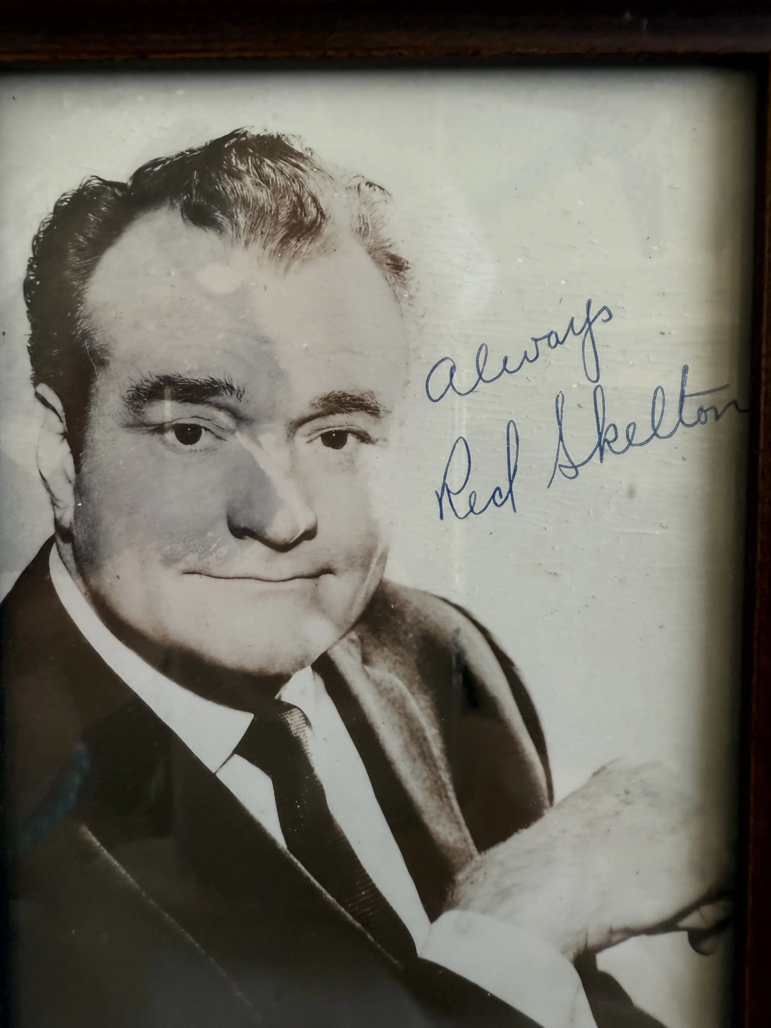 Black and white photograph of a man with neatly styled hair, wearing a suit and tie, with handwritten signature 'Always, Red Skelton' in blue ink on the right side.
