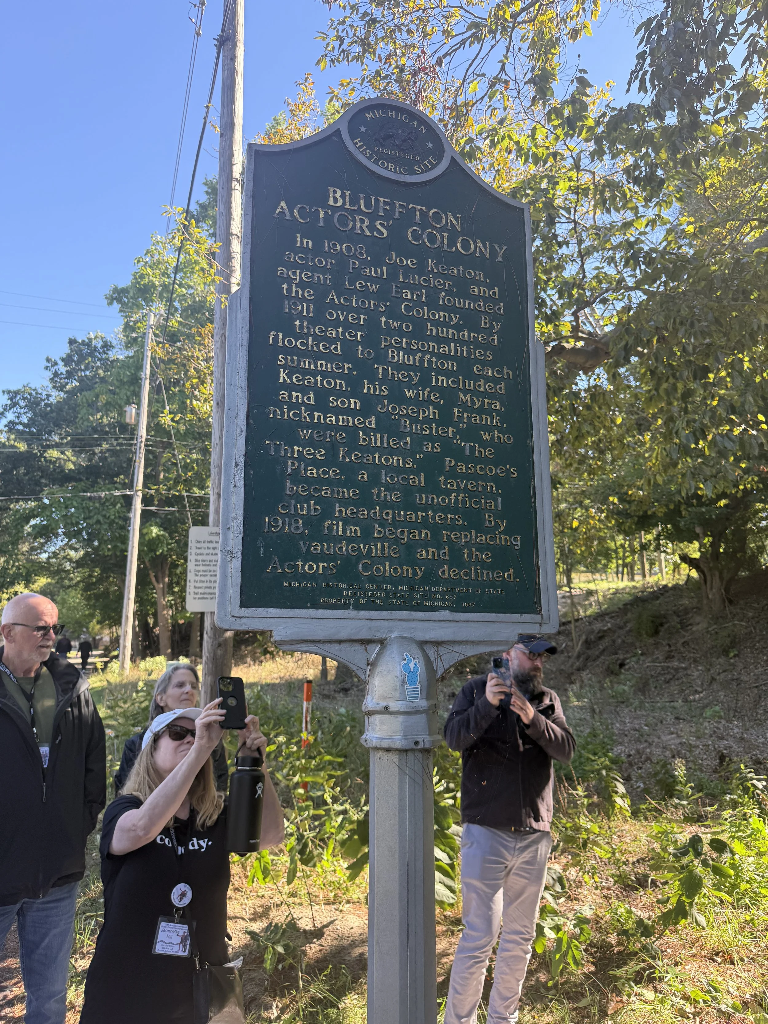 Historical marker sign about the Blufton Actors' Colony, with people standing around taking photos in a park-like setting with trees and clear blue sky in the background.