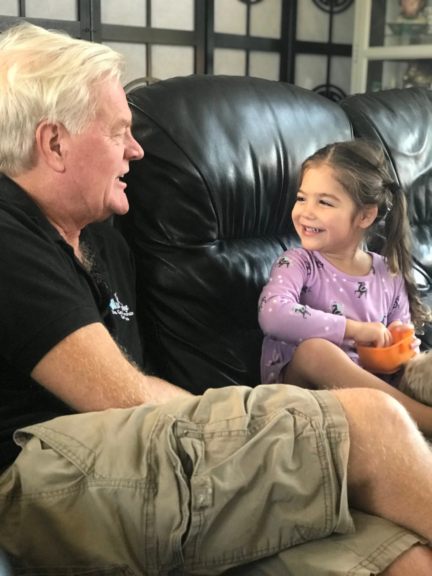 A man with gray hair and a young girl with brown hair sitting on a black leather couch, smiling and looking at each other.