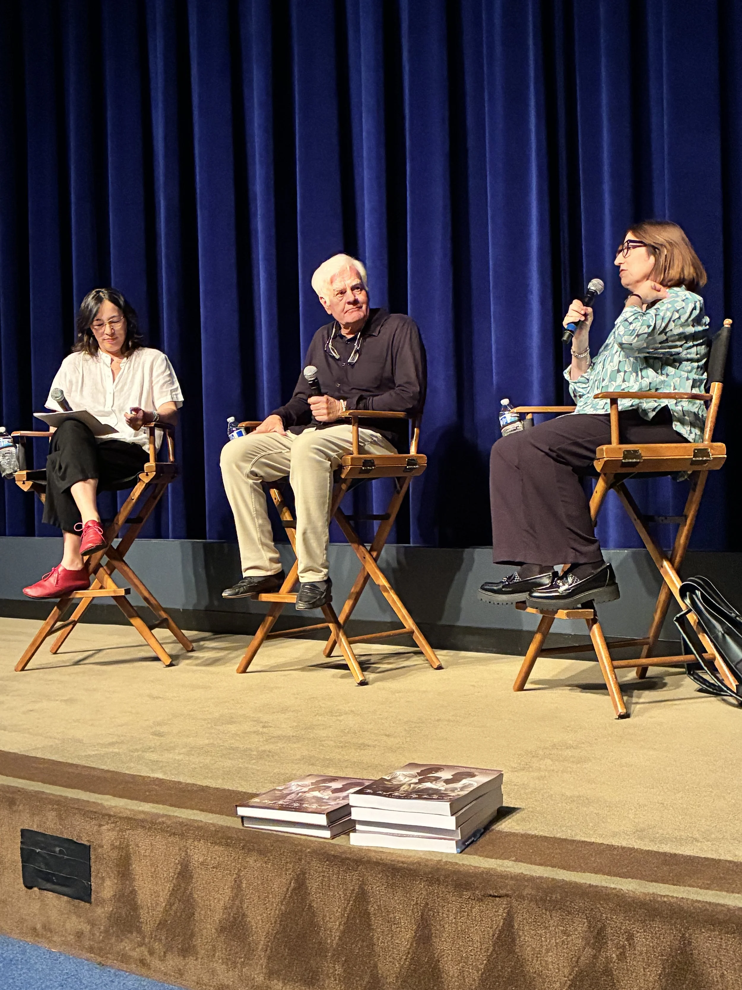Three women seated on a stage with a blue curtain backdrop, engaged in a discussion. One woman is holding a microphone and wearing glasses. Two books are stacked on the front of the stage.