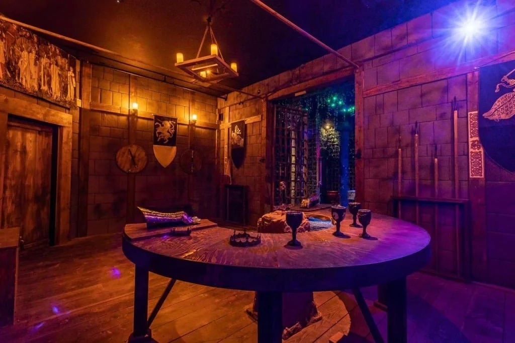 A medieval-style room with wooden walls, medieval shields, and banners. There is a round wooden table with goblets and a crown, lit by candles; an ornate chandelier hangs from the ceiling. A window with lattice and foliage outside is visible.