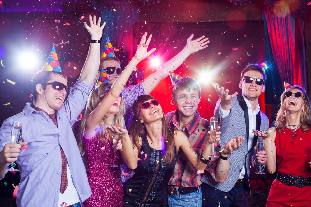 People wearing party hats and sunglasses celebrating with confetti and champagne in a lively party setting.