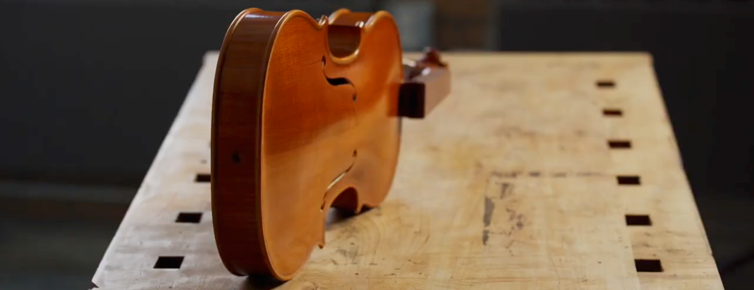 A frame from the North Bennet Street School Portrait Project - A small wooden violin body placed upright on a wooden workbench in a workshop.