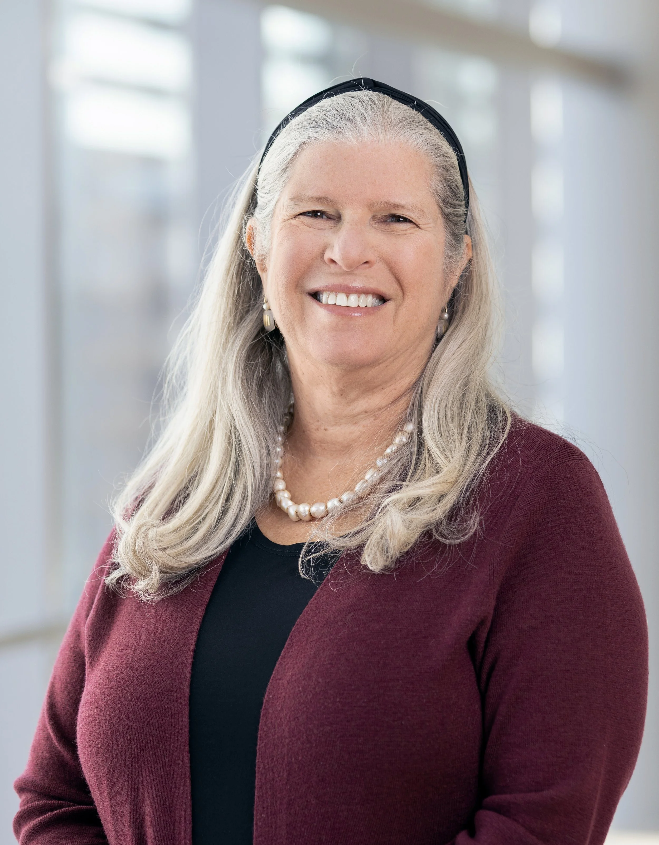 A smiling older woman with long gray hair, wearing a dark top, a pearl necklace, pearl earrings, and a burgundy blazer. She is standing indoors with a bright, blurred background of windows.