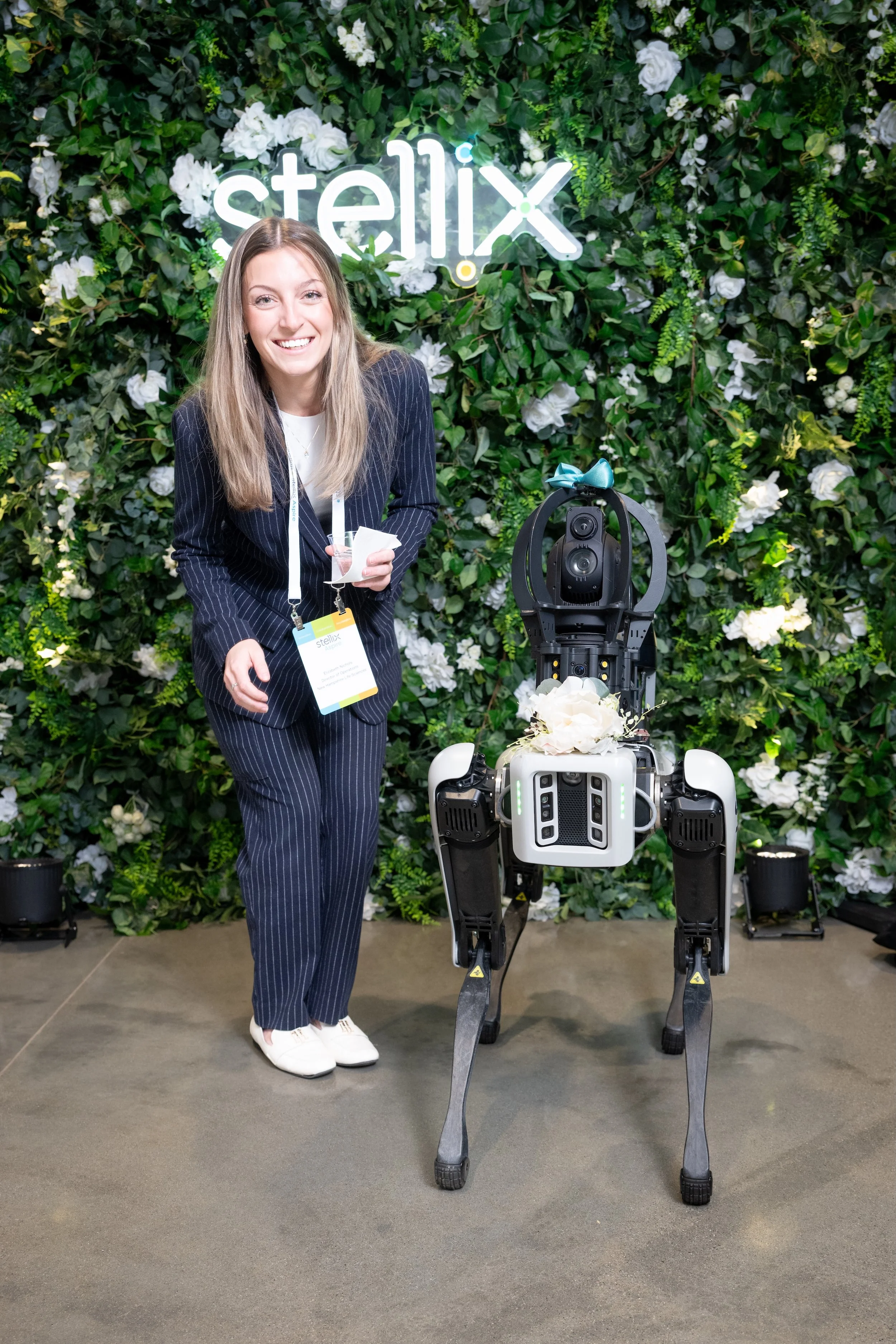 A woman with long blonde hair smiling at a technology exhibition, standing in front of a green wall with white flowers and a neon sign that says 'Stellix', with a Boston Dynamics robot dog in front of her decorated with a blue bow.