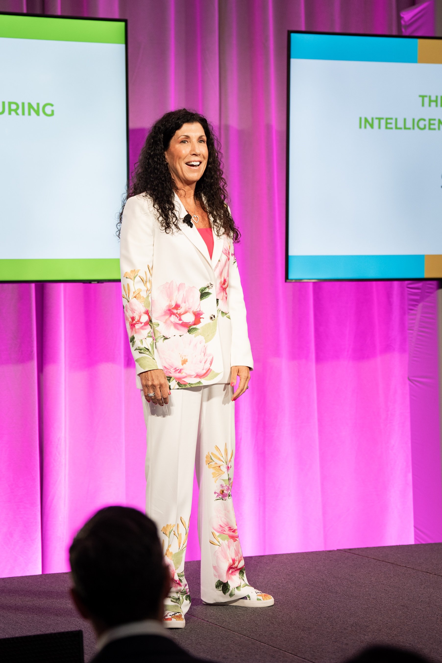 A woman with dark curly hair wearing a white blazer with floral patterns and matching floral pants stands on stage in front of television screens, speaking to an audience at a conference or presentation.