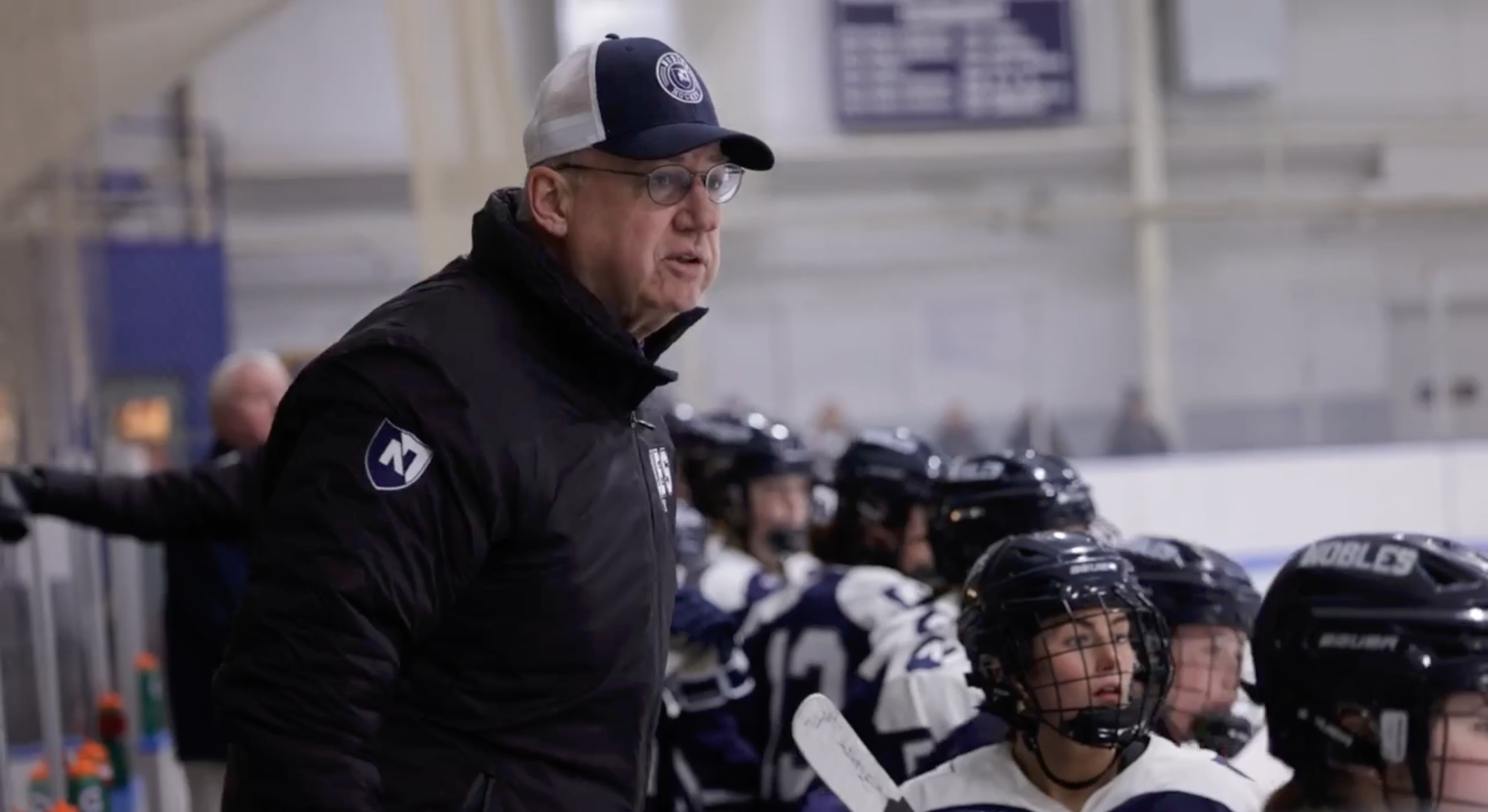 A frame from the Noble and Greenough Coach Resor Retirement Video - a coach (Tom Resor) speaking to ice hockey players on the bench during a game inside an ice rink.