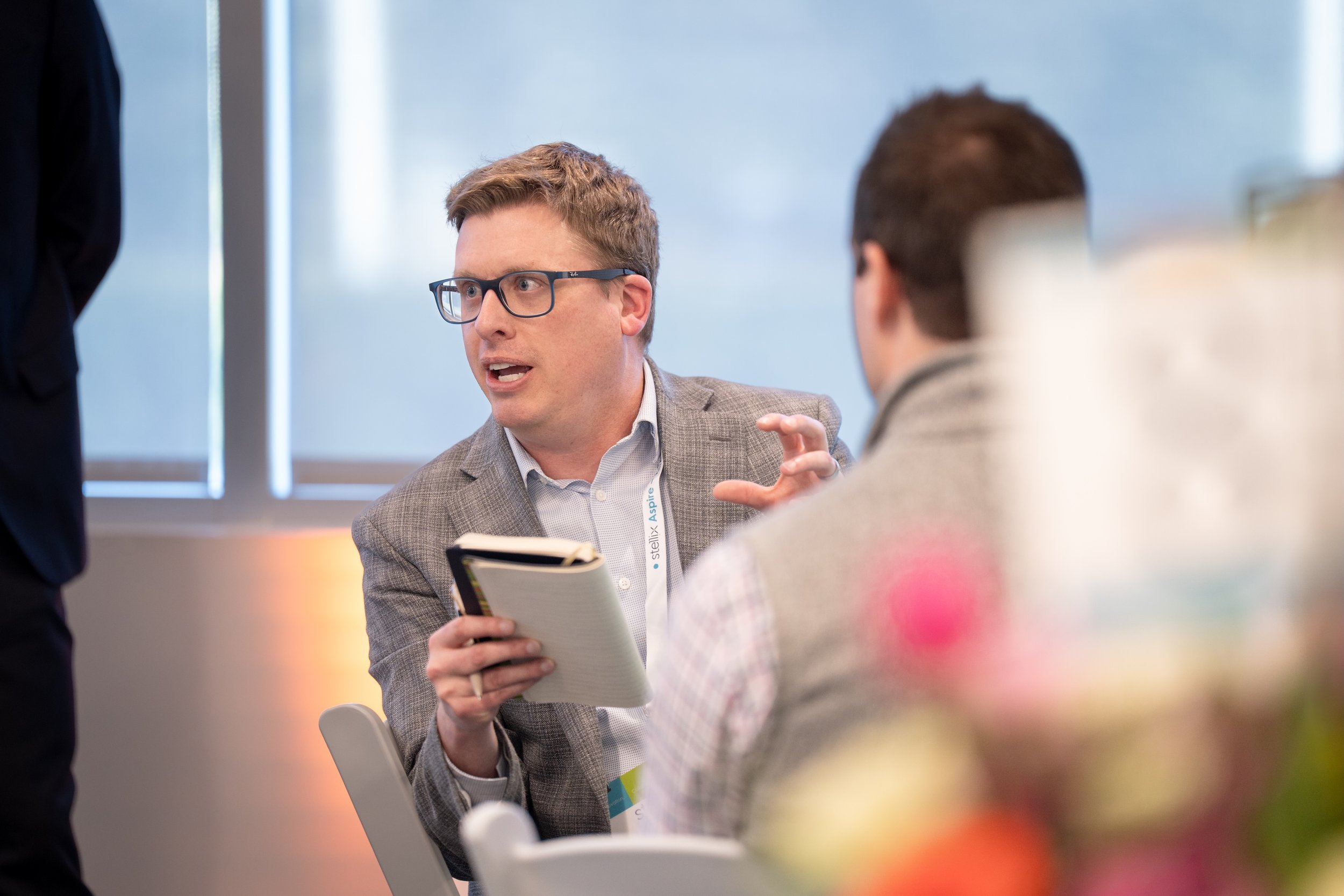 A man with glasses speaking and gesturing while holding a notebook in a professional setting during a meeting or discussion.