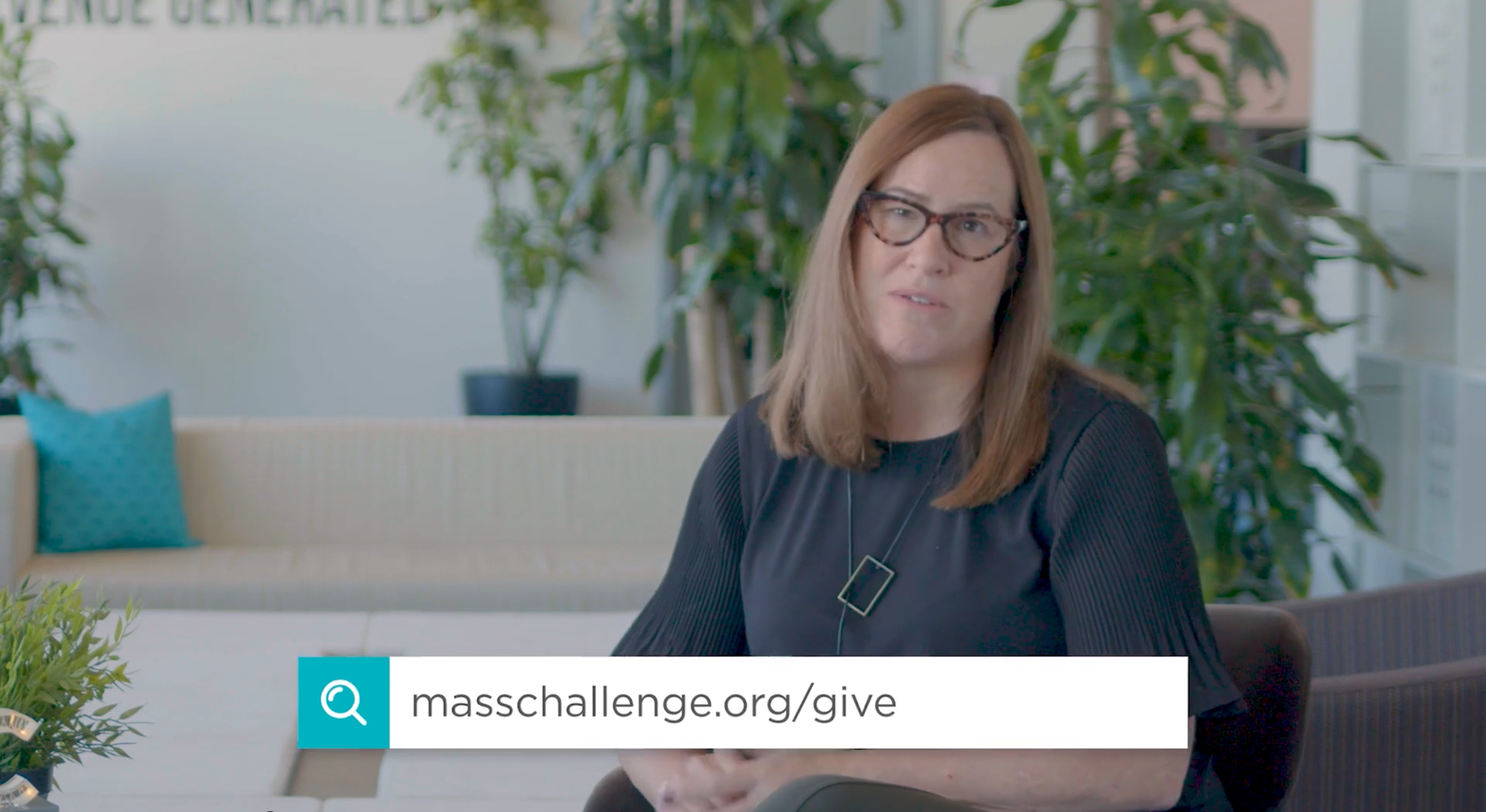 A frame from the MassChallenge remote gala - A woman - the CEO of MassChallenge - with brown hair, wearing glasses and a black top, sitting indoors in front of green plants, with a couch in the background, looking at the camera.