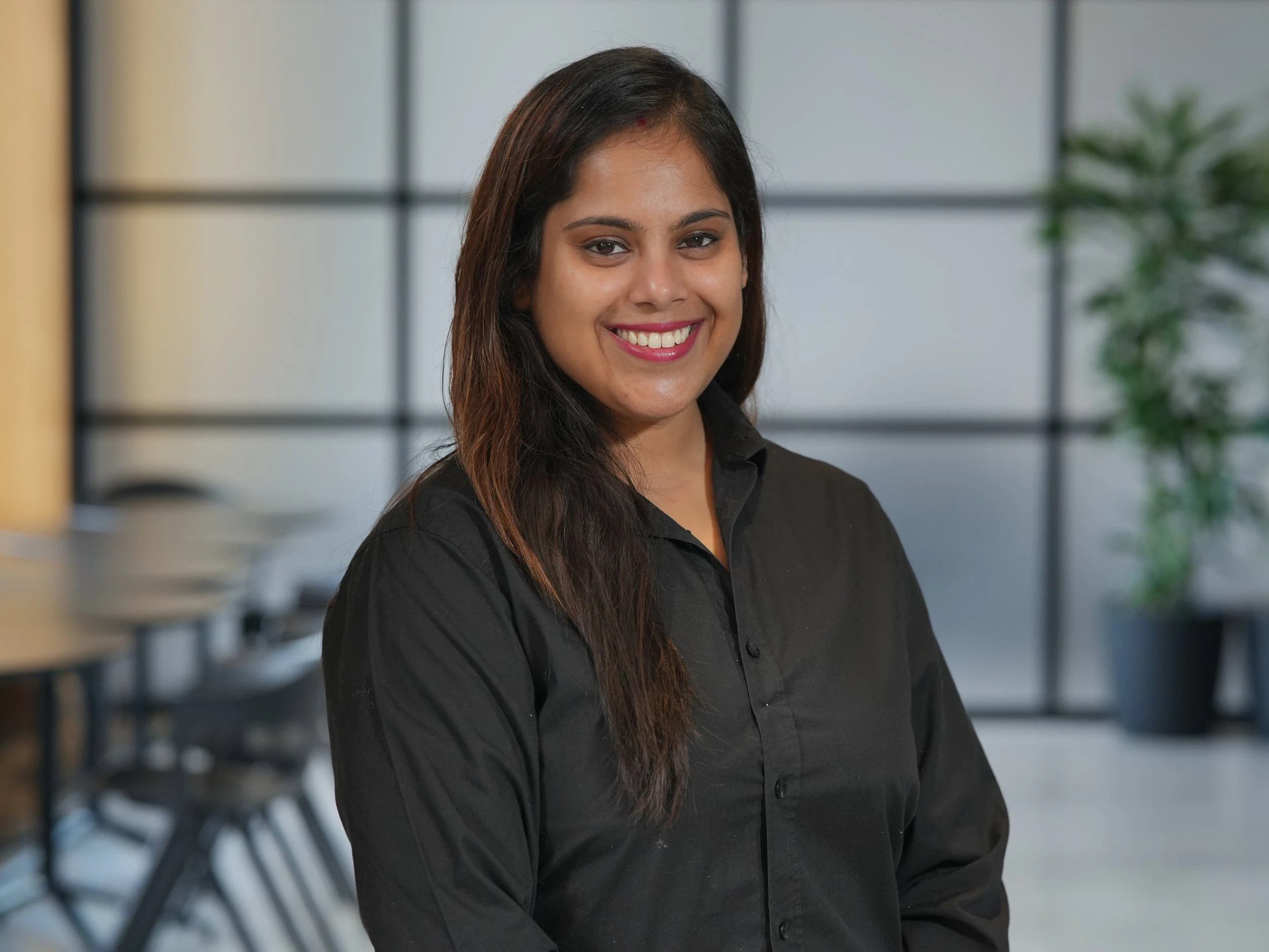 A woman with long dark hair wearing a black blouse, smiling, in a modern office setting with chairs and green plants in the background.