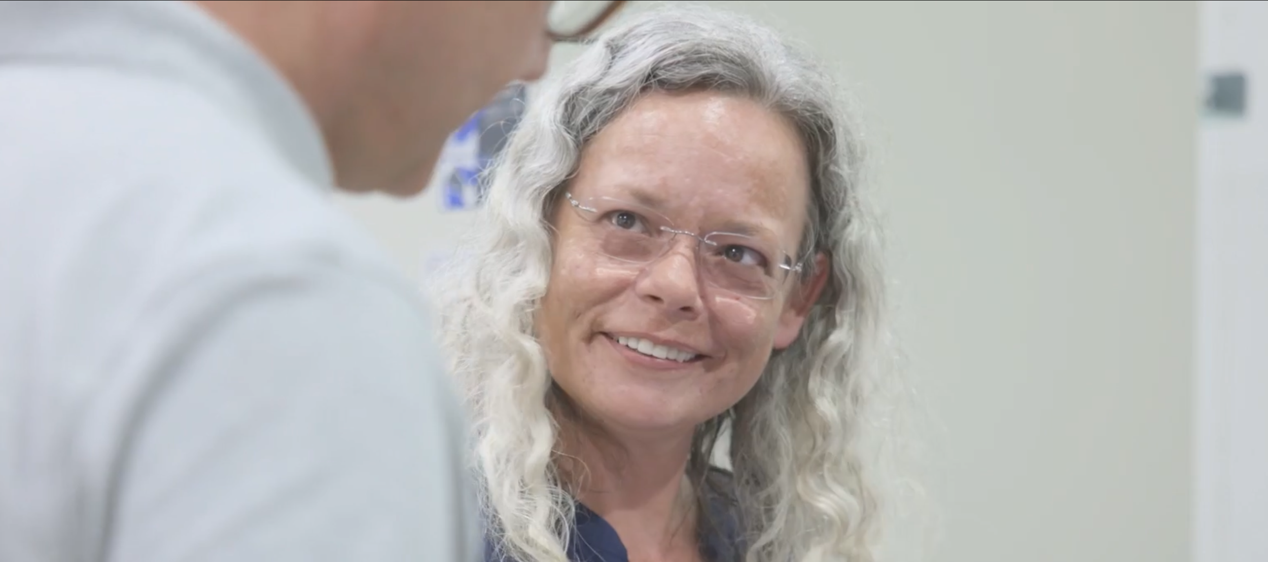 A frame from the ProKidney videos - a woman with gray, curly hair and glasses smiles at a colleague, in a clinical setting.