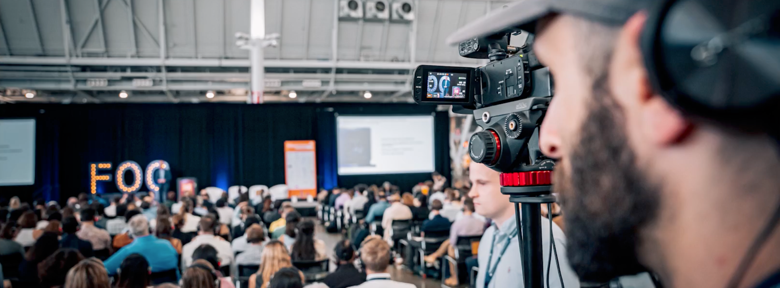 A camera operator - Andy - operating a professional video camera at a conference a large audience and stage in the background.