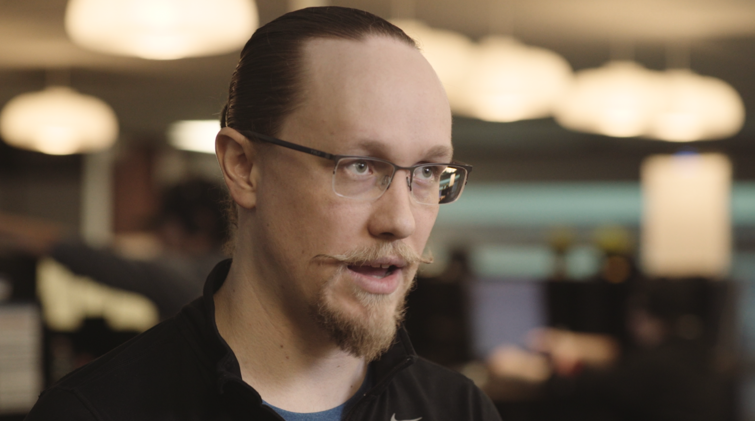 A frame from the New Voice Media Testimonial video - a subject, with glasses, red hair, and a beard, speaking to an off-camera interviewer, with blurred lights in an office in the background.