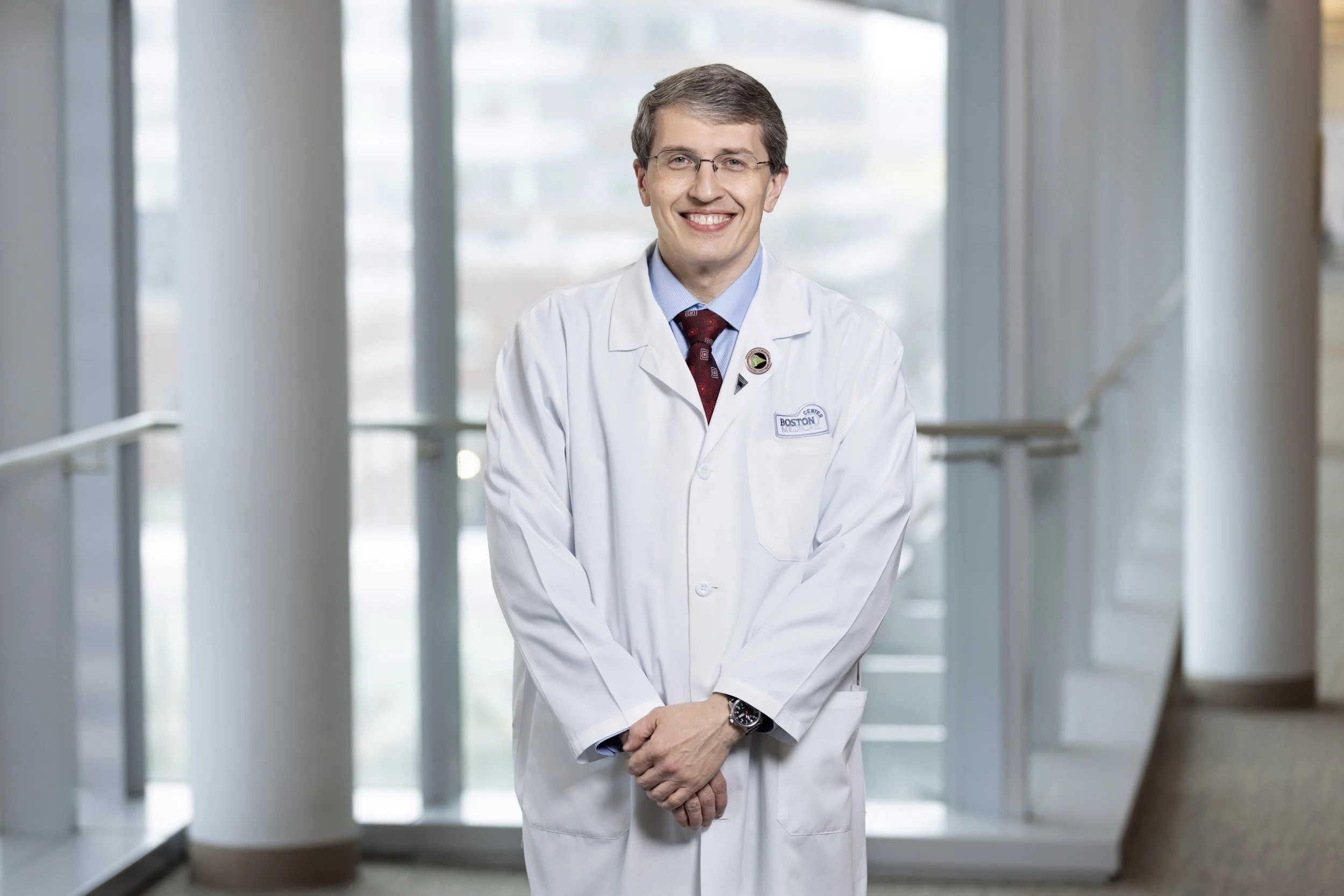 A smiling male doctor wearing glasses, a white lab coat with a badge, a blue shirt, and a patterned maroon tie, standing in a bright, modern hospital corridor with large windows.