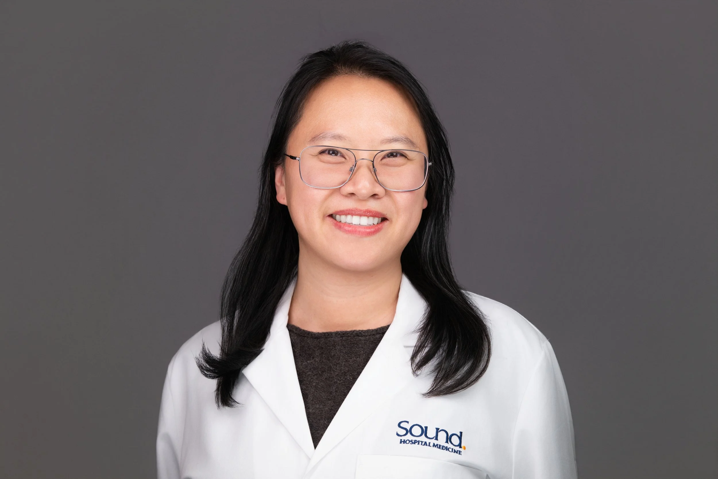 Portrait /headshot of an Asian woman with long black hair, wearing glasses, a white coat with 'Sound Hospital Medicine' logo, smiling against a gray background.