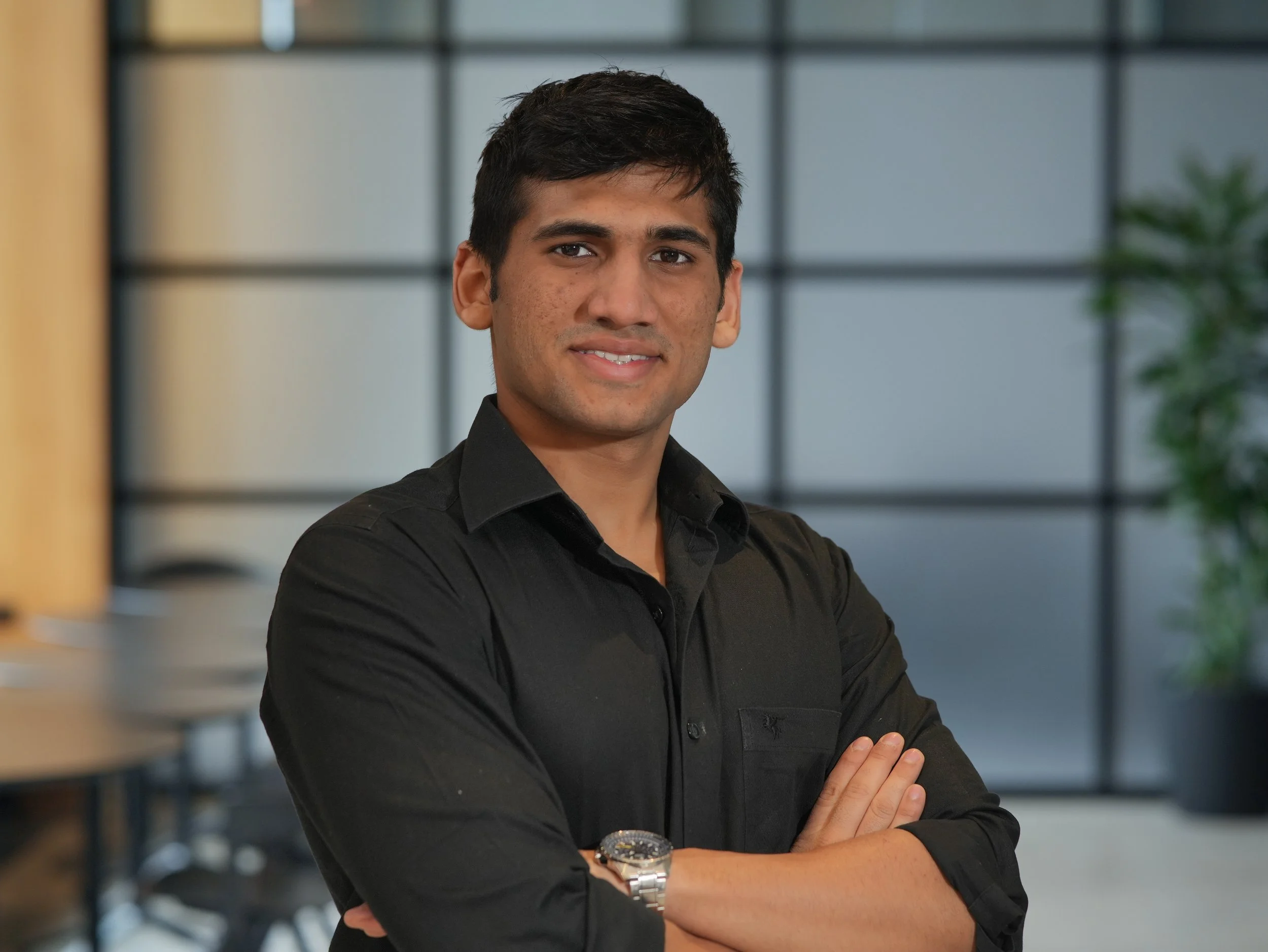 A young man with short dark hair and medium brown skin, wearing a black shirt and a silver watch, standing with arms crossed in front of a modern office background with large windows and a potted plant.