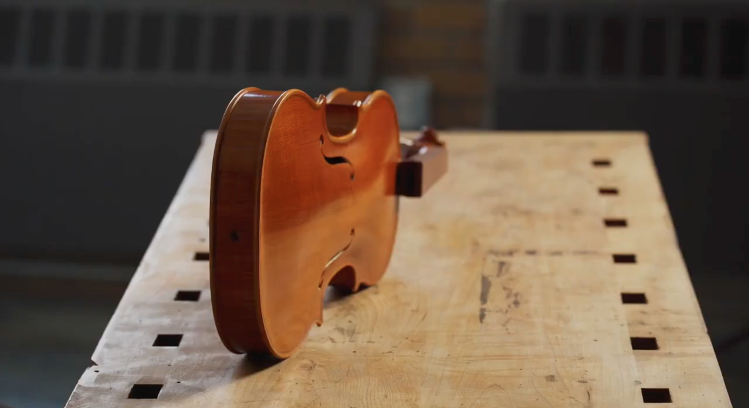 A frame from the North Bennet Street School Portrait Project - A small wooden violin body placed upright on a wooden workbench in a workshop.