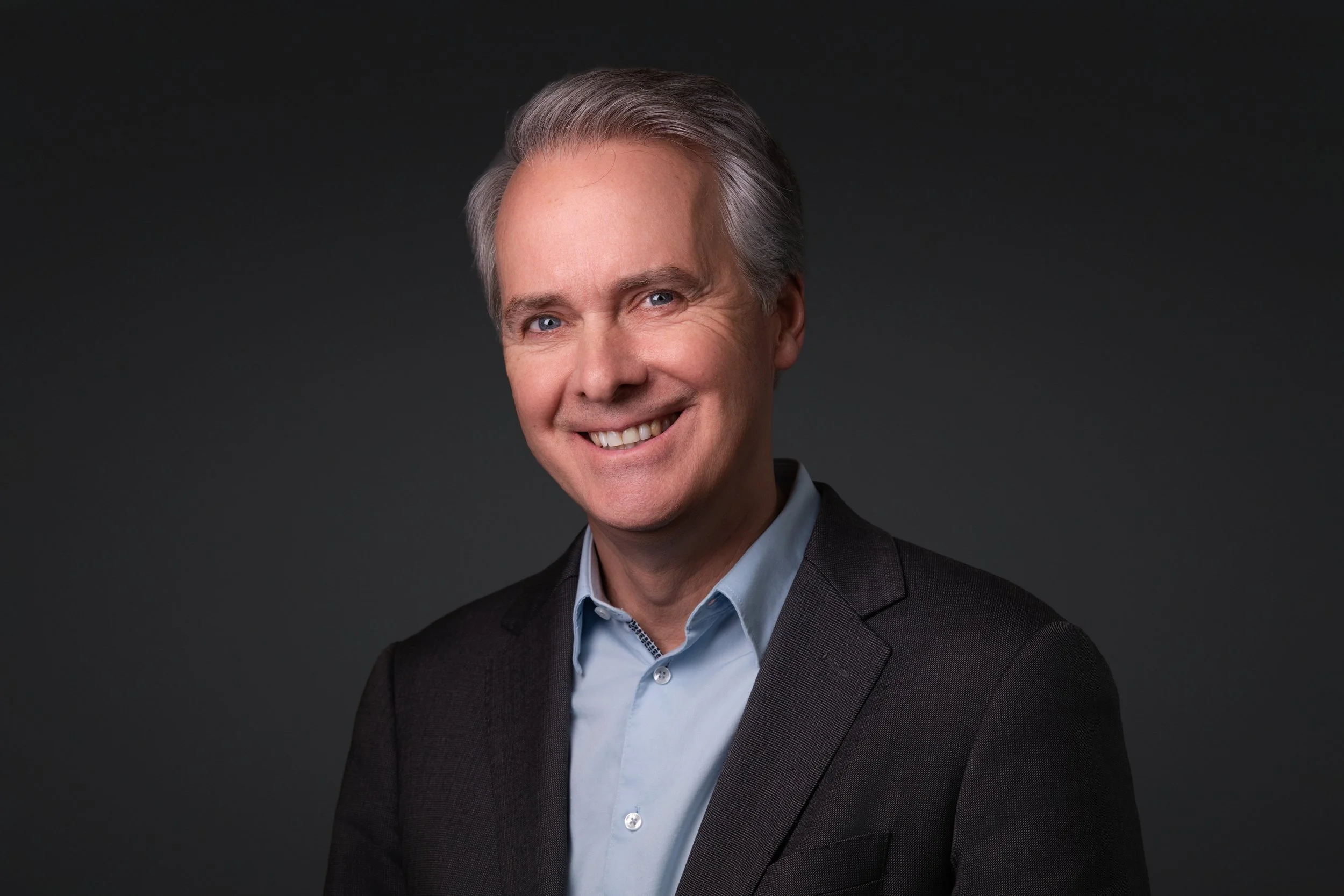 Portrait/headshot of a professional middle-aged man with gray hair and blue eyes, smiling, wearing a dark suit jacket and light blue shirt, against a dark background.