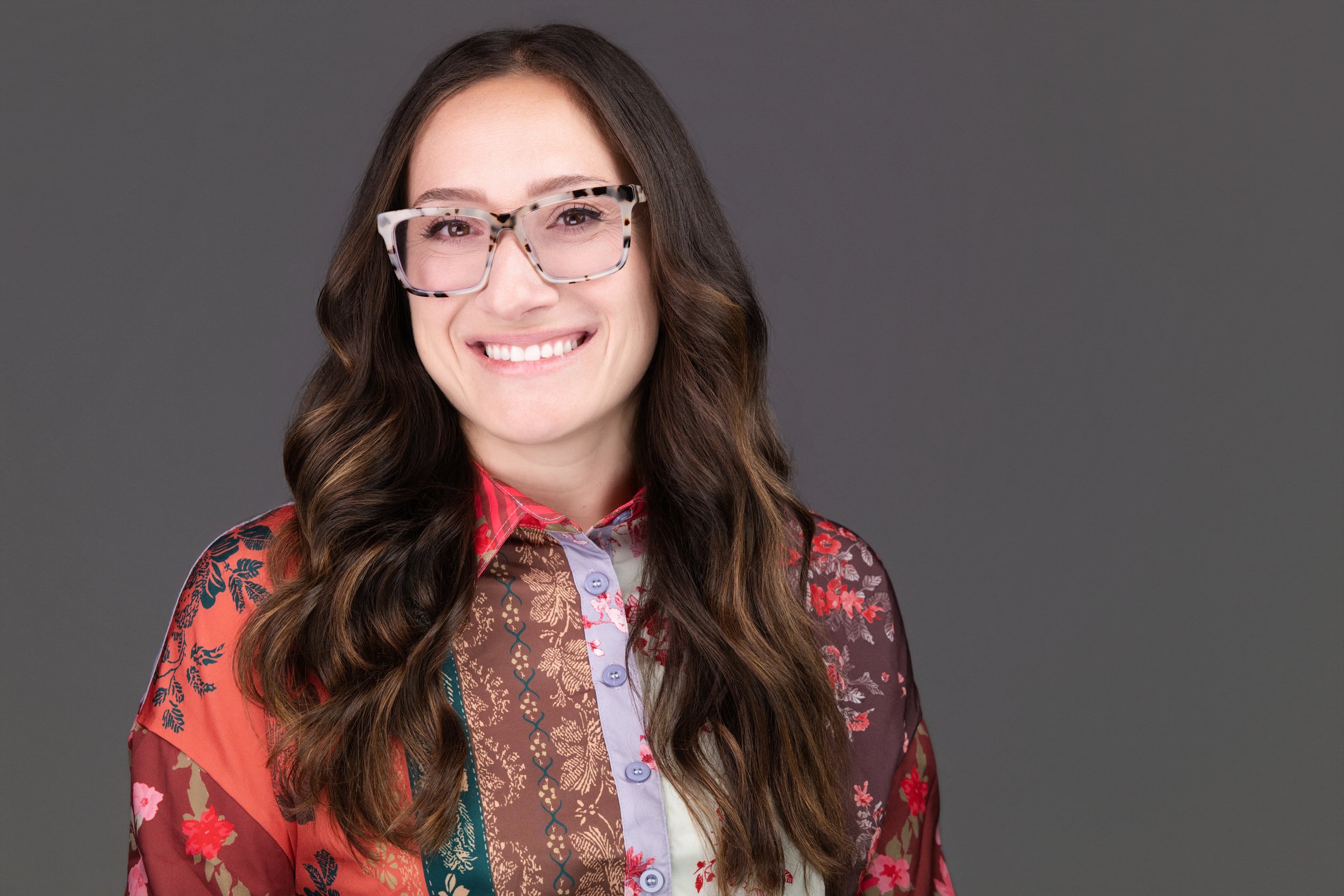 Portrait/headshot of a woman with long wavy brown hair smiling, wearing large patterned glasses, a multicolored patchwork shirt, and a floral outer layer against a plain gray background.