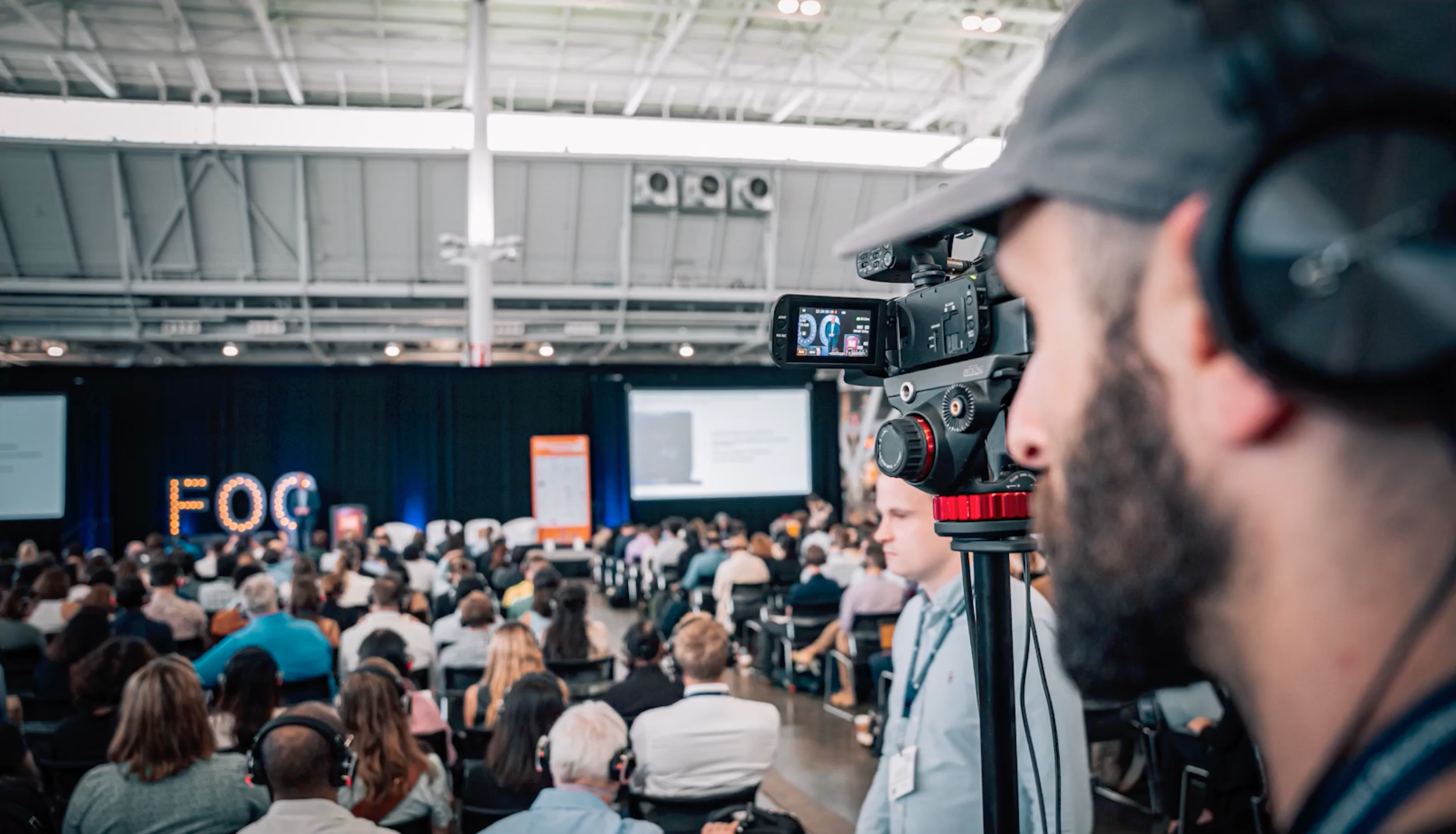 A camera operator - Andy - operating a professional video camera at a conference a large audience and stage in the background.