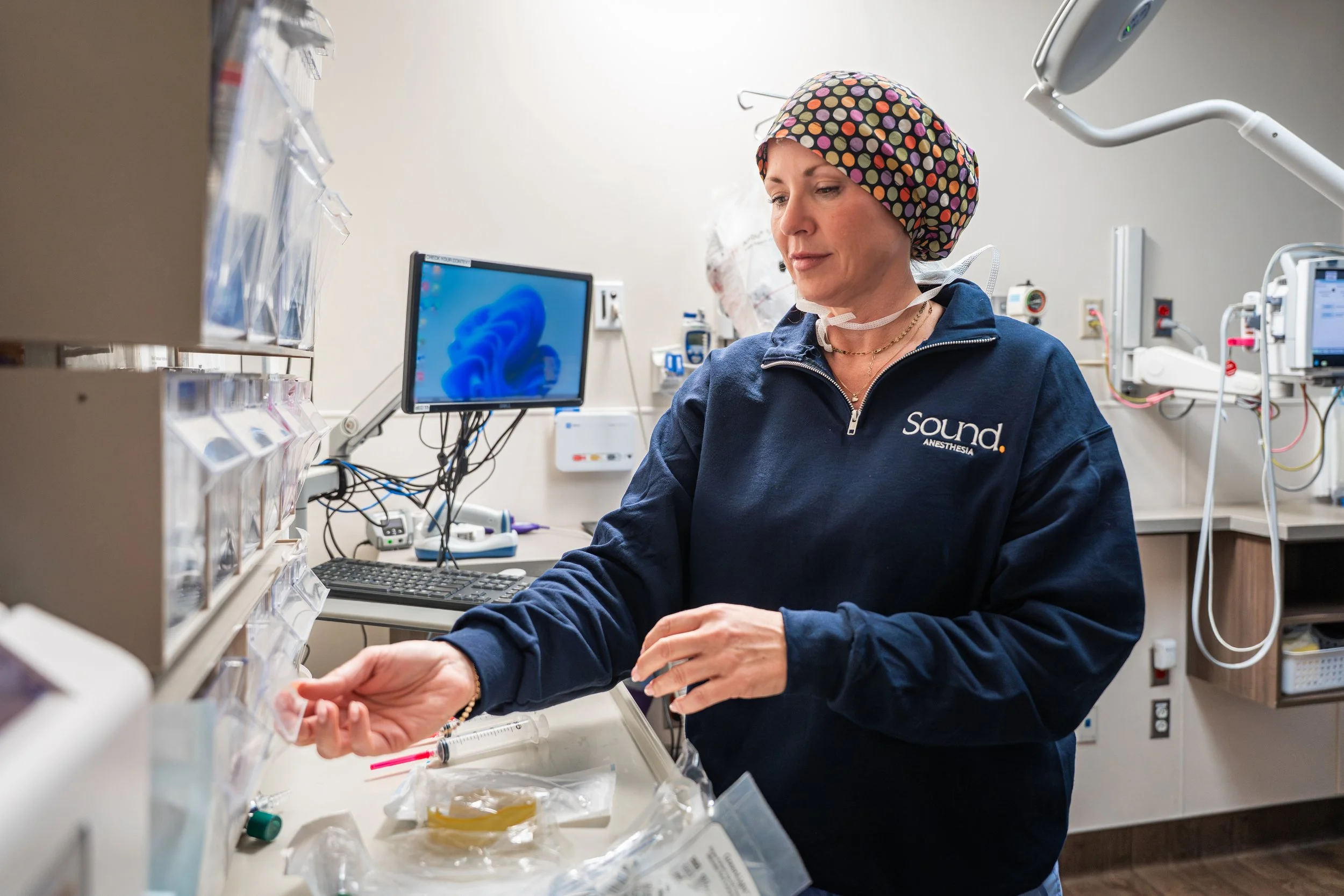 A woman wearing a colorful polka dot surgical cap and navy sweatshirt with 'Sound Anesthesia' is in a medical setting, preparing medication or supplies with medical equipment and monitors in the background.