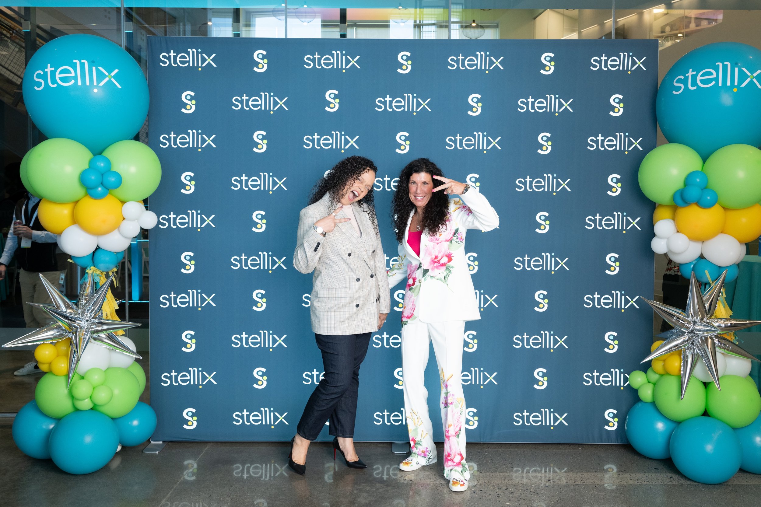 Two women posing happily in front of a blue step-and-repeat backdrop with 'Stellix' logos, surrounded by colorful balloons and silver star-shaped balloons at a promotional event.