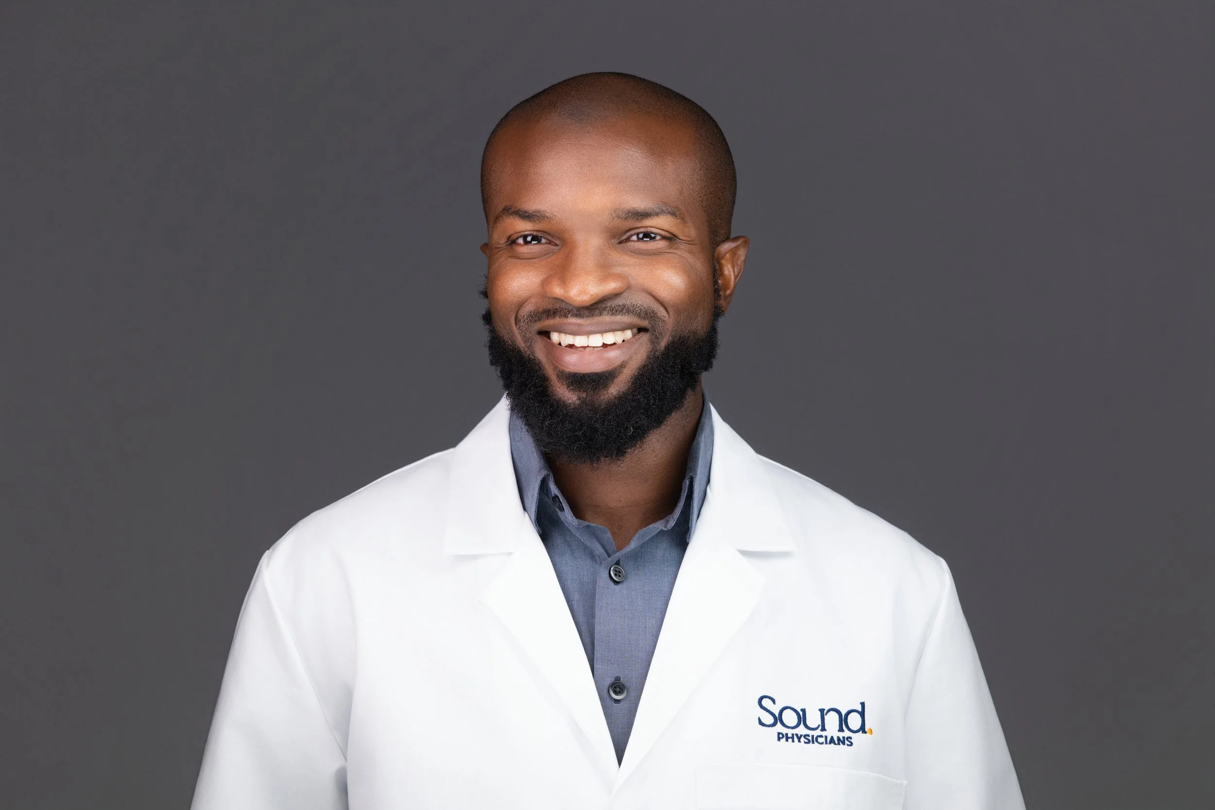 Portrait /headshot of a smiling African American male doctor in a white coat with 'Sound Physicians' logo, dark grey background.
