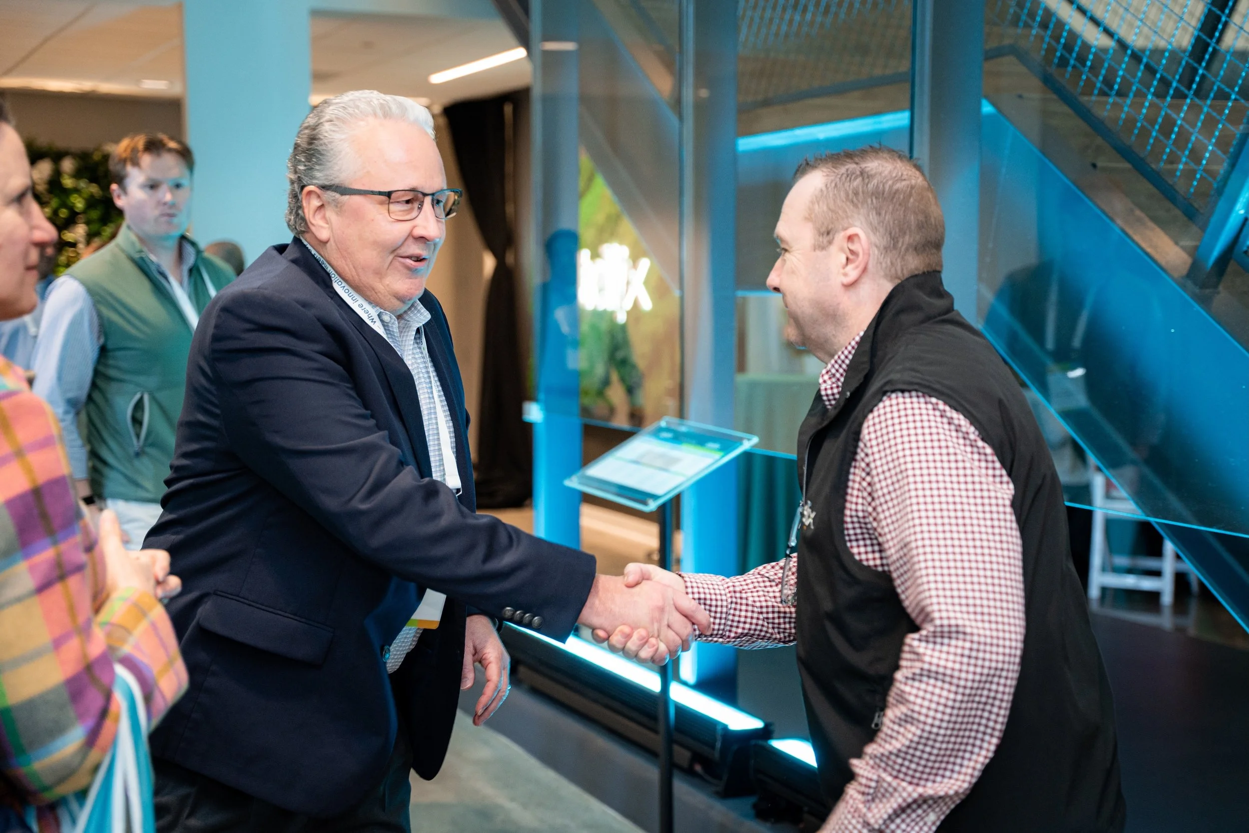 Two men in business attire shaking hands at a professional event, with other people in the background and a blue illuminated display in an indoor setting.
