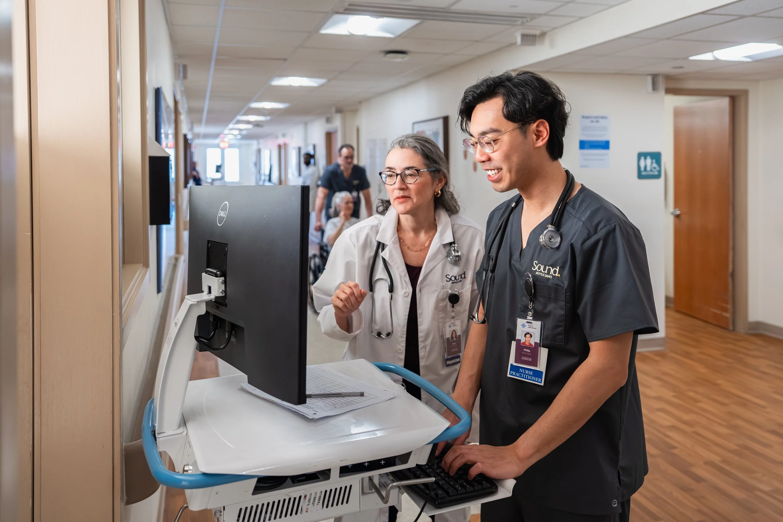 Medical professionals, a woman and a young man, standing in a hospital corridor near a computer station, both wearing stethoscopes and identification badges, smiling and looking at a computer screen.