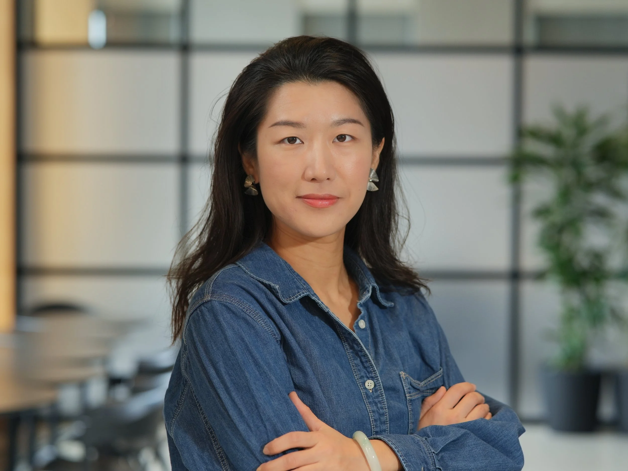 A woman with dark hair wearing a denim shirt and silver earrings standing indoors with arms crossed, against a background of shelves and a plant.