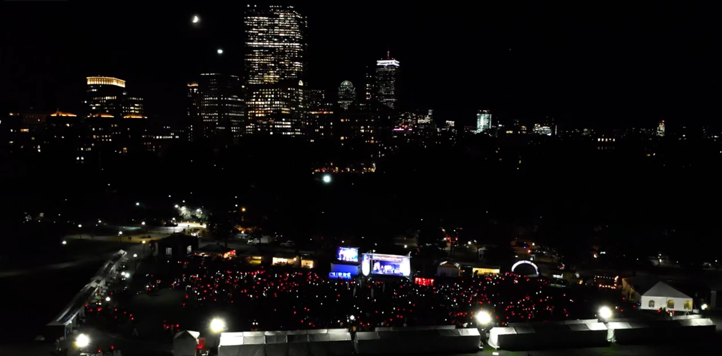 A frame from the Syndax Pharmaceuticals Light the Night videos - nighttime Boston skyline with many tall illuminated skyscrapers, visible moon, and a large crowd holding red, yellow, and white lights, for the Leukemia and Lymphoma Society.