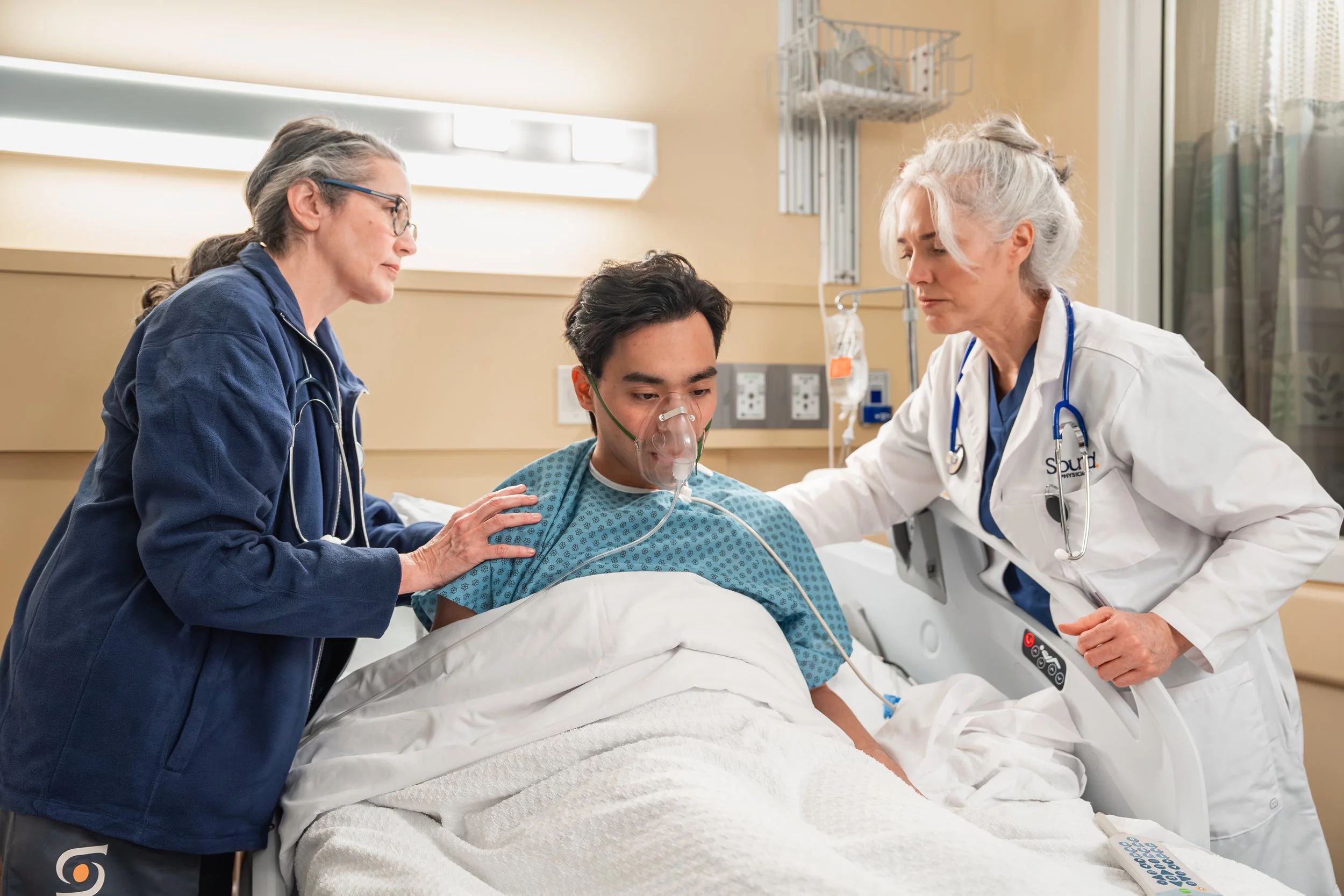 Medical team attending patient in hospital bed, one woman in blue scrubs with stethoscope, another woman in white coat with stethoscope, man with oxygen mask, hospital room setting.