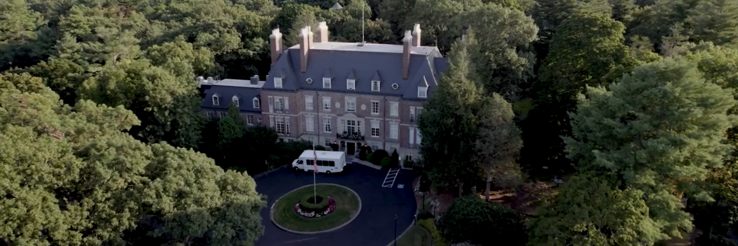 A frame from the Ruderman Executive Summit video - aerial view from a drone of a large historic mansion surrounded by trees, with a circular driveway, flagpole, and a white vehicle parked near the entrance.