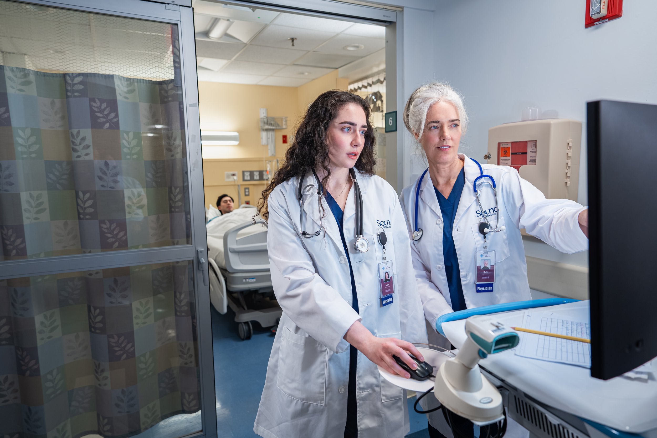 Two female healthcare professionals, both wearing white lab coats with identification badges, are working together in a hospital or medical setting. They are focused on a computer or monitor, with one using a computer mouse. A hospital bed with a patient in the background is visible, along with medical equipment and hospital room features.