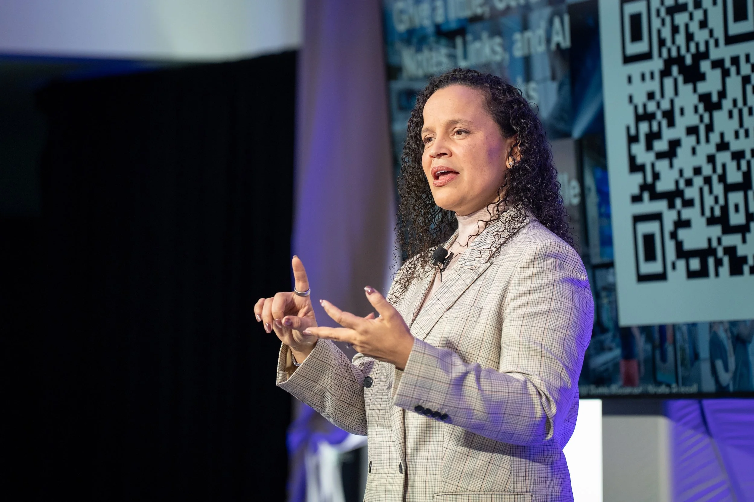 A woman with dark, curly hair wearing a beige checkered blazer is speaking at a conference. She is gesturing with her hands. Behind her, there is a large screen displaying a QR code and some text.