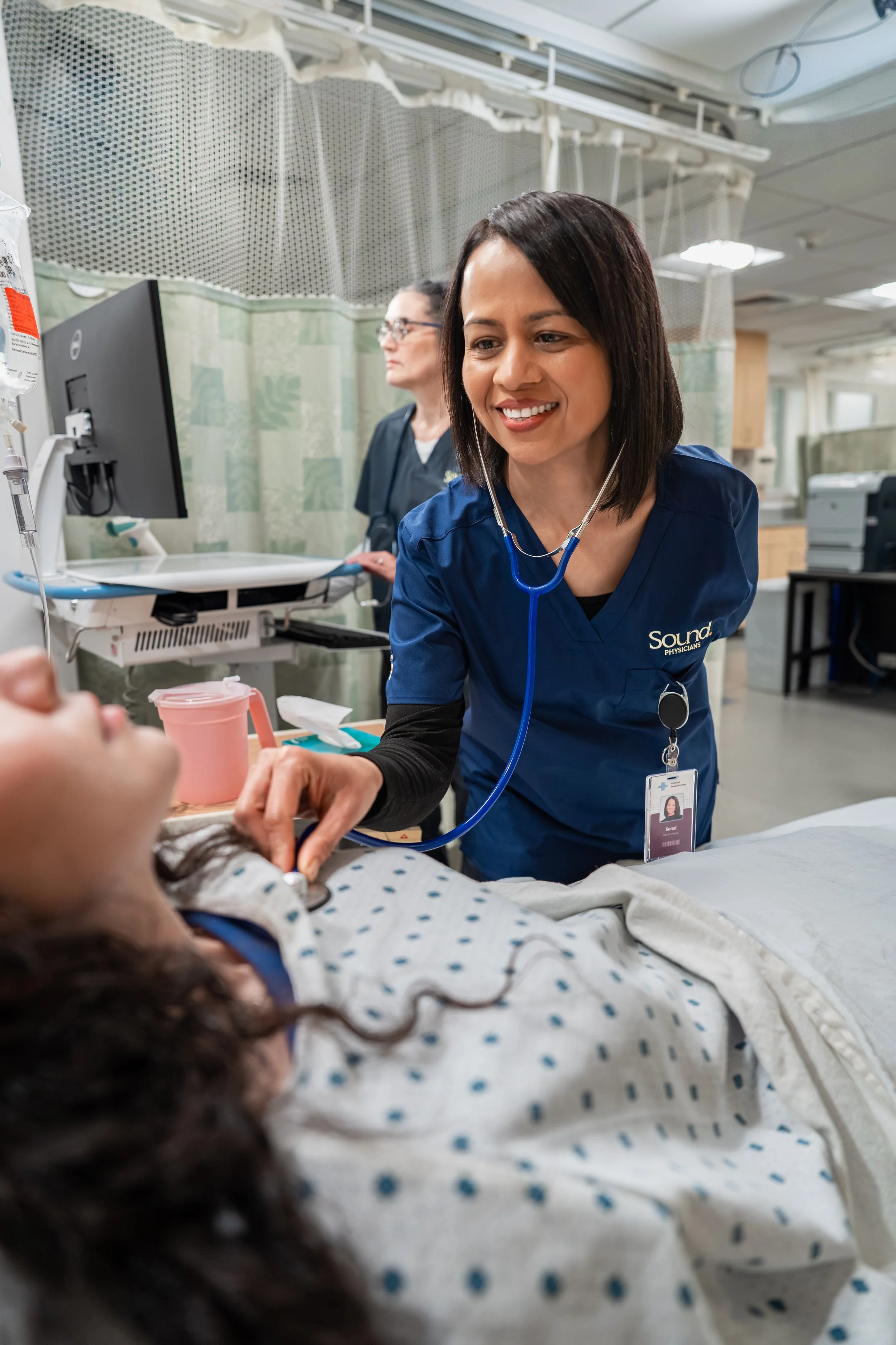 A healthcare professional in blue scrubs with a stethoscope around her neck is smiling as she examines a patient lying in a hospital bed. The patient appears to be a young woman with long, dark hair, wearing a hospital gown, lying on her back.