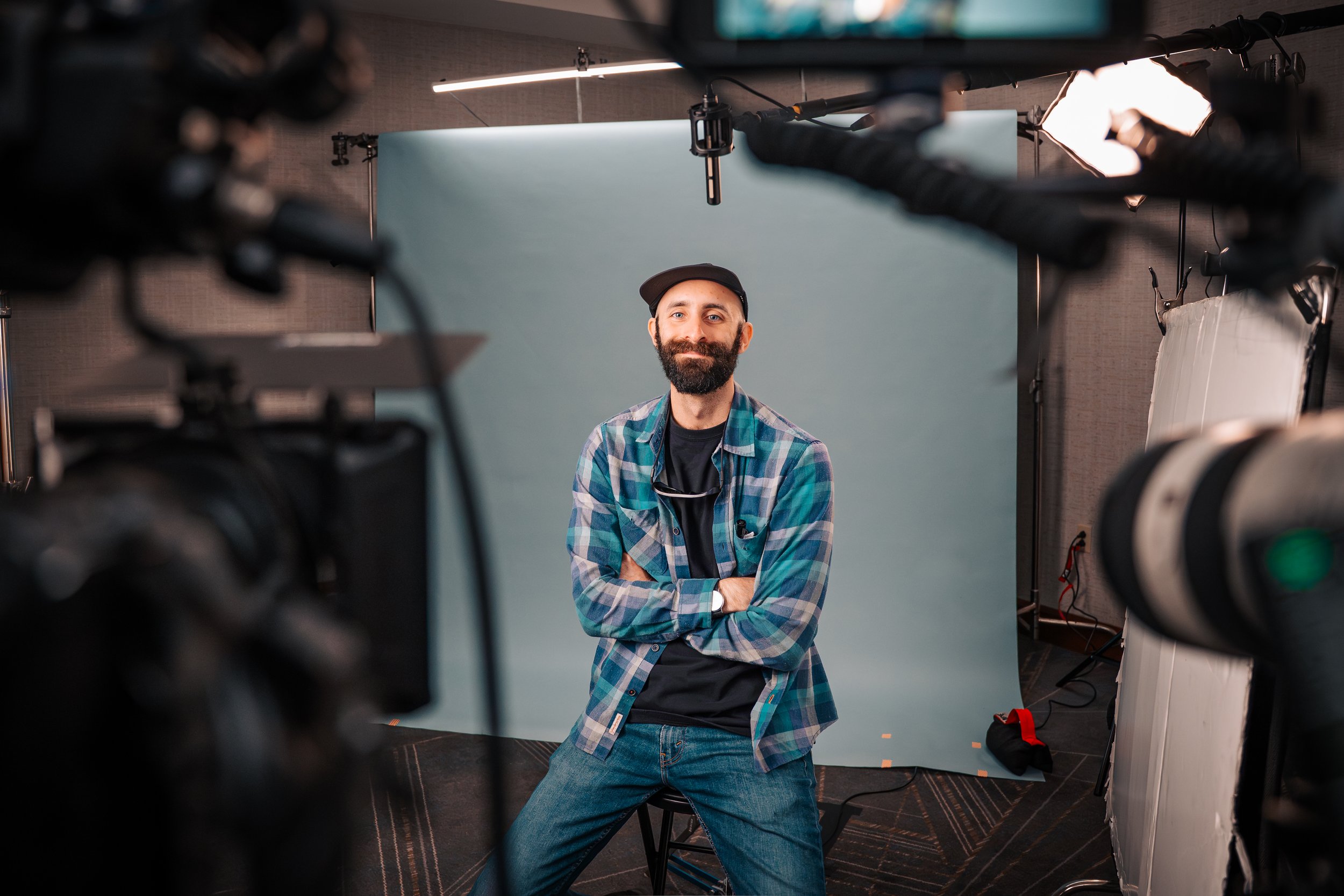 A man with a beard - Andy - wearing a plaid shirt and a black baseball cap. He is seated with his arms crossed smiling at the cameras in a studio surrounded by lighting equipment.