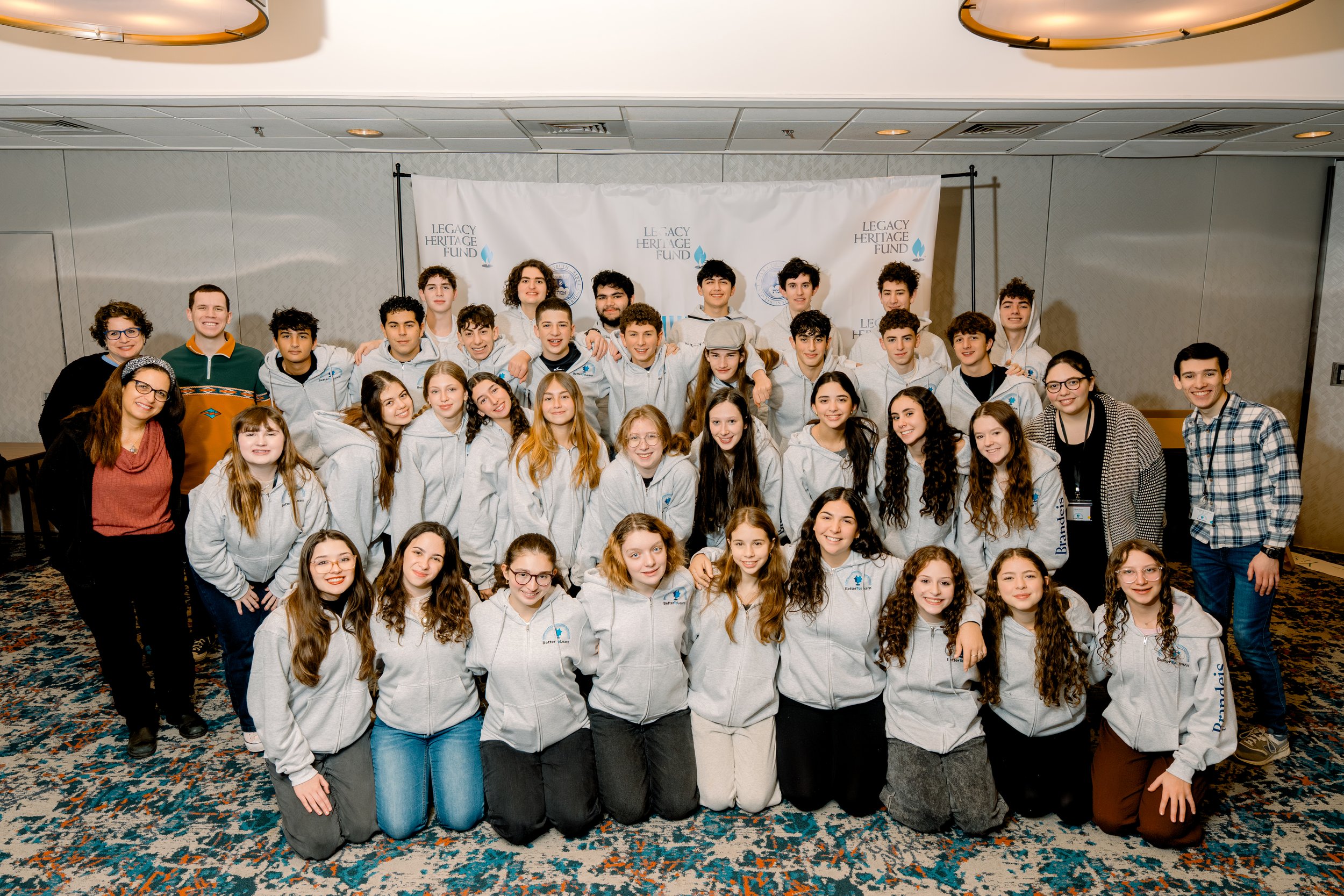 A large group of young people and adults posing together in front of a banner that says 'Legacy Heritage Fund'. The group includes mostly teenagers wearing gray hoodies with a logo, and some adults, in a room with patterned carpet and ceiling lights.