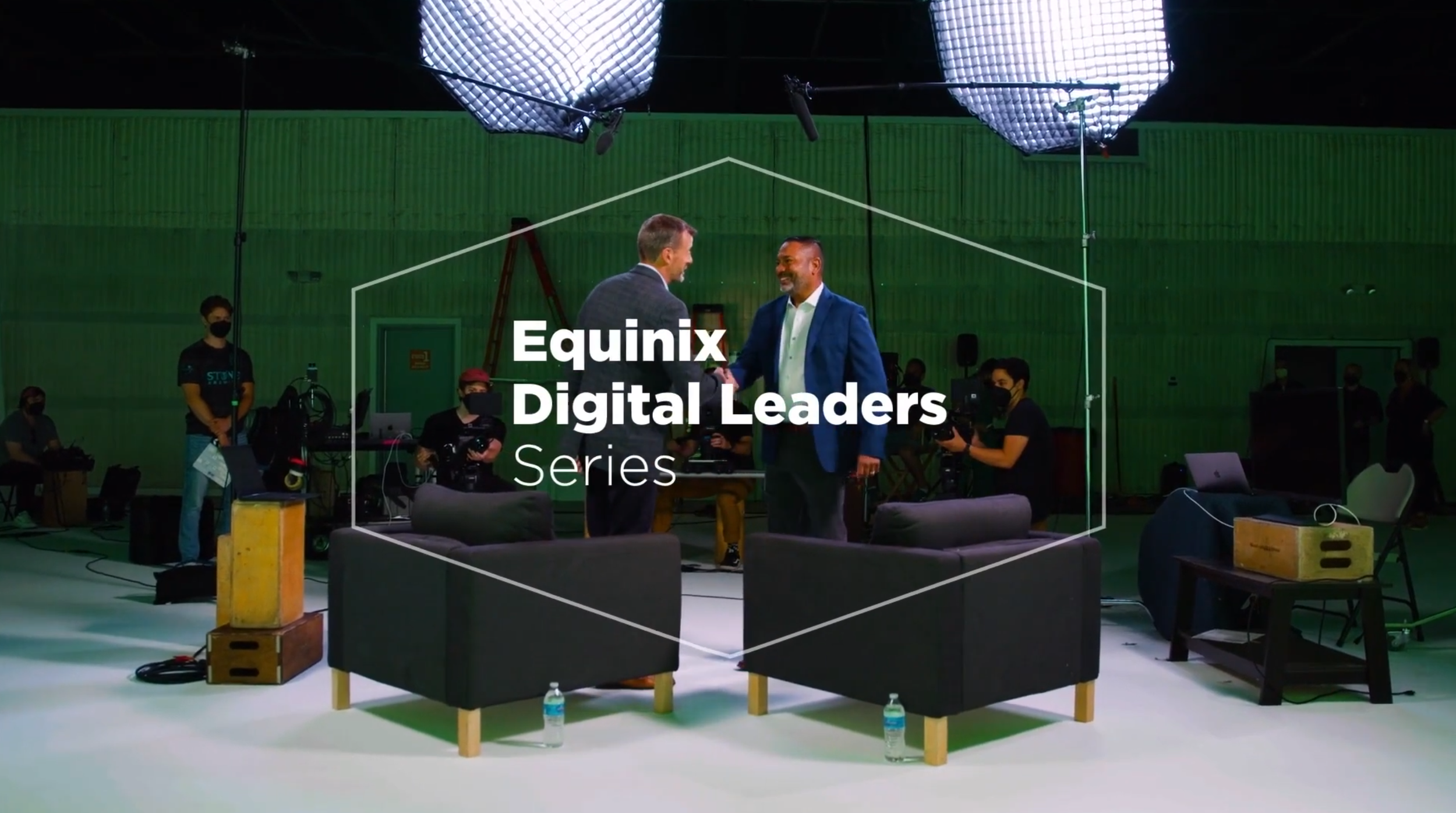 A frame from the Equinix Digital Leaders Series - Two men shaking hands in a film studio with lighting equipment, surrounded by seated camera operators with cameras and computers. The text on the image reads 'Equinix Digital Leaders Series.'