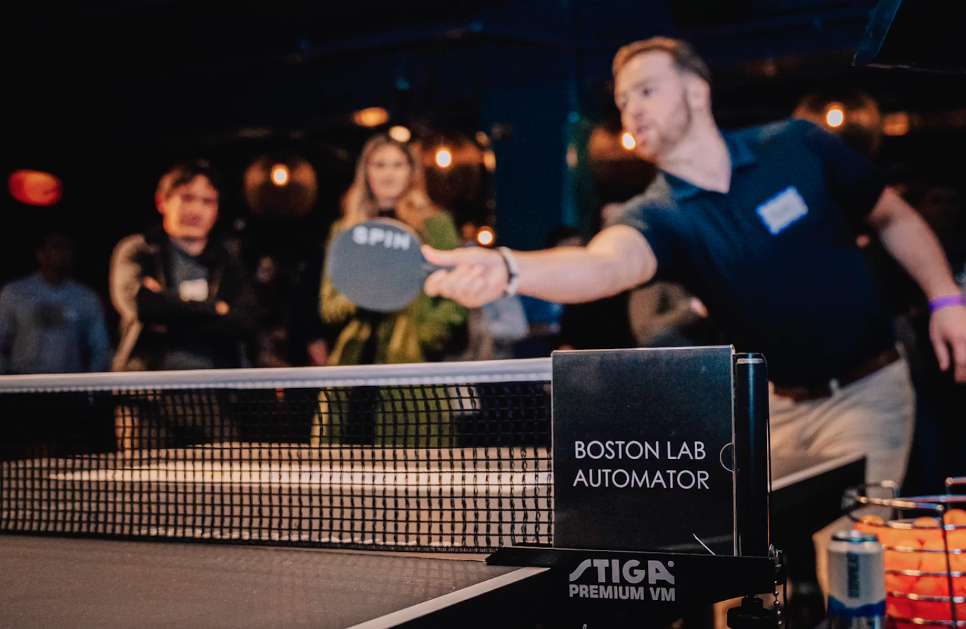 A frame from the Boston Lab Automators video - a man playing ping pong at an event, with people watching in the background. The ping pong paddle has the word 'SPIN' printed on it. There is a sign that reads 'BOSTON LAB AUTOMATOR'.